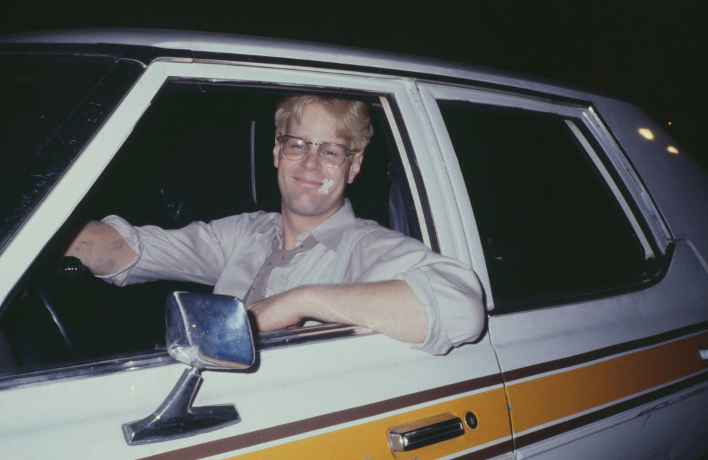 A man with glasses and a smile sits in the driver's seat of an older-model car, looking at the camera