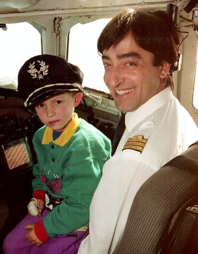 A child sits on the lap of a smiling airplane pilot in a cockpit. The child wears a pilot's cap and a colorful sweater with a cartoon character