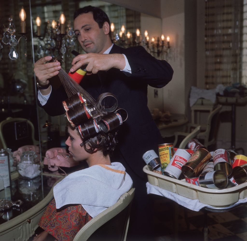A stylist uses rollers on a seated woman's hair in a vintage salon, surrounded by styling products and tools