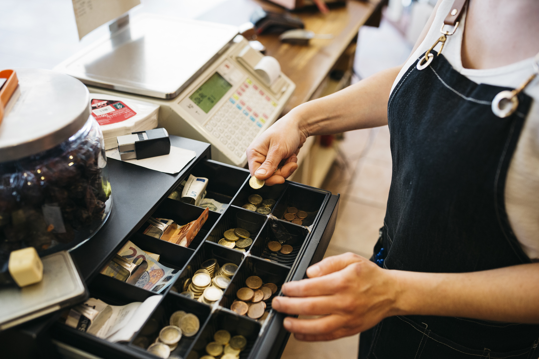 A person wearing an apron is organizing coins and bills in a cash register drawer at a store counter, with a jar and register visible