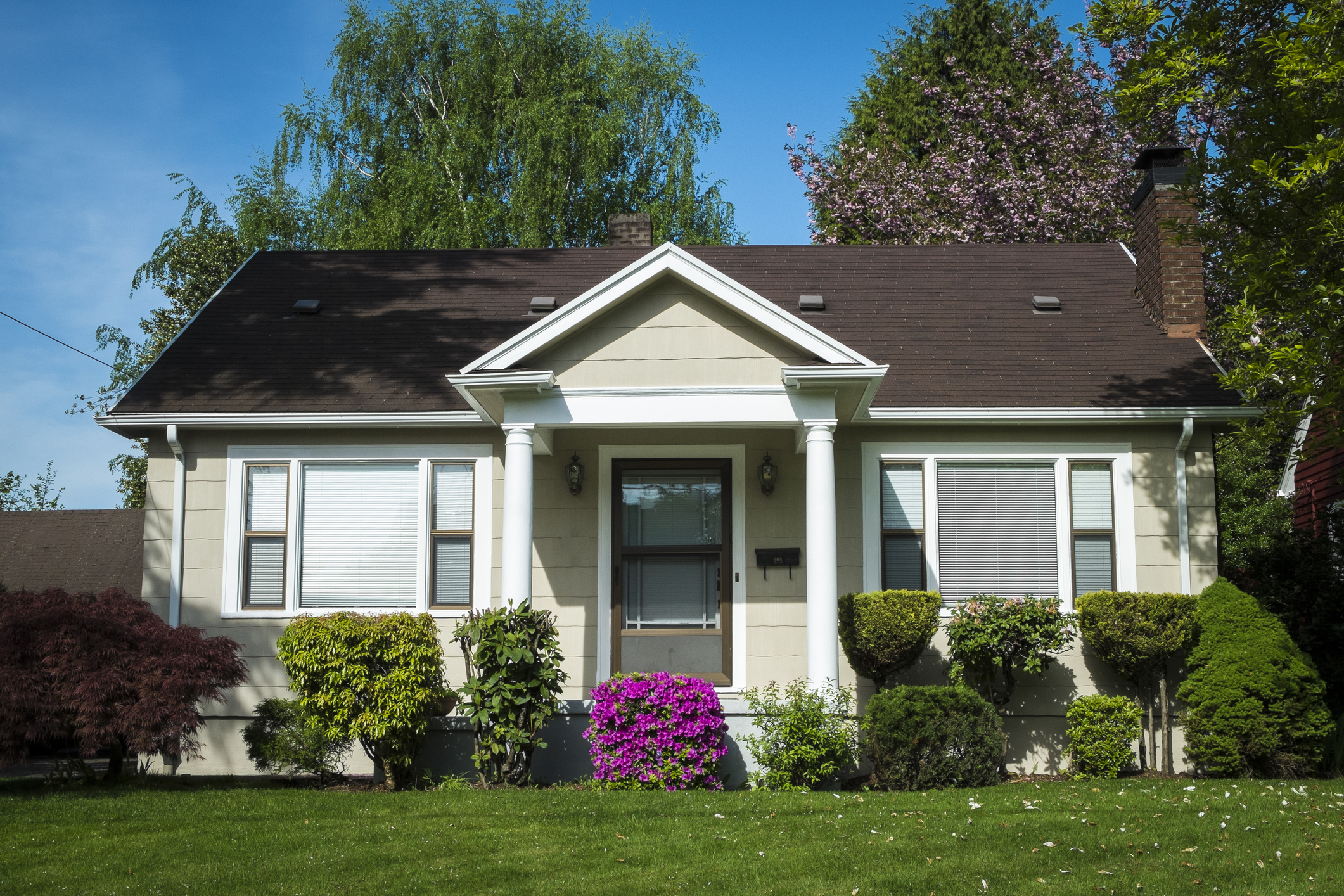 A quaint single-story house with a well-maintained front yard, large bushes, and a blooming shrub near the entrance. The house has a front porch and large windows