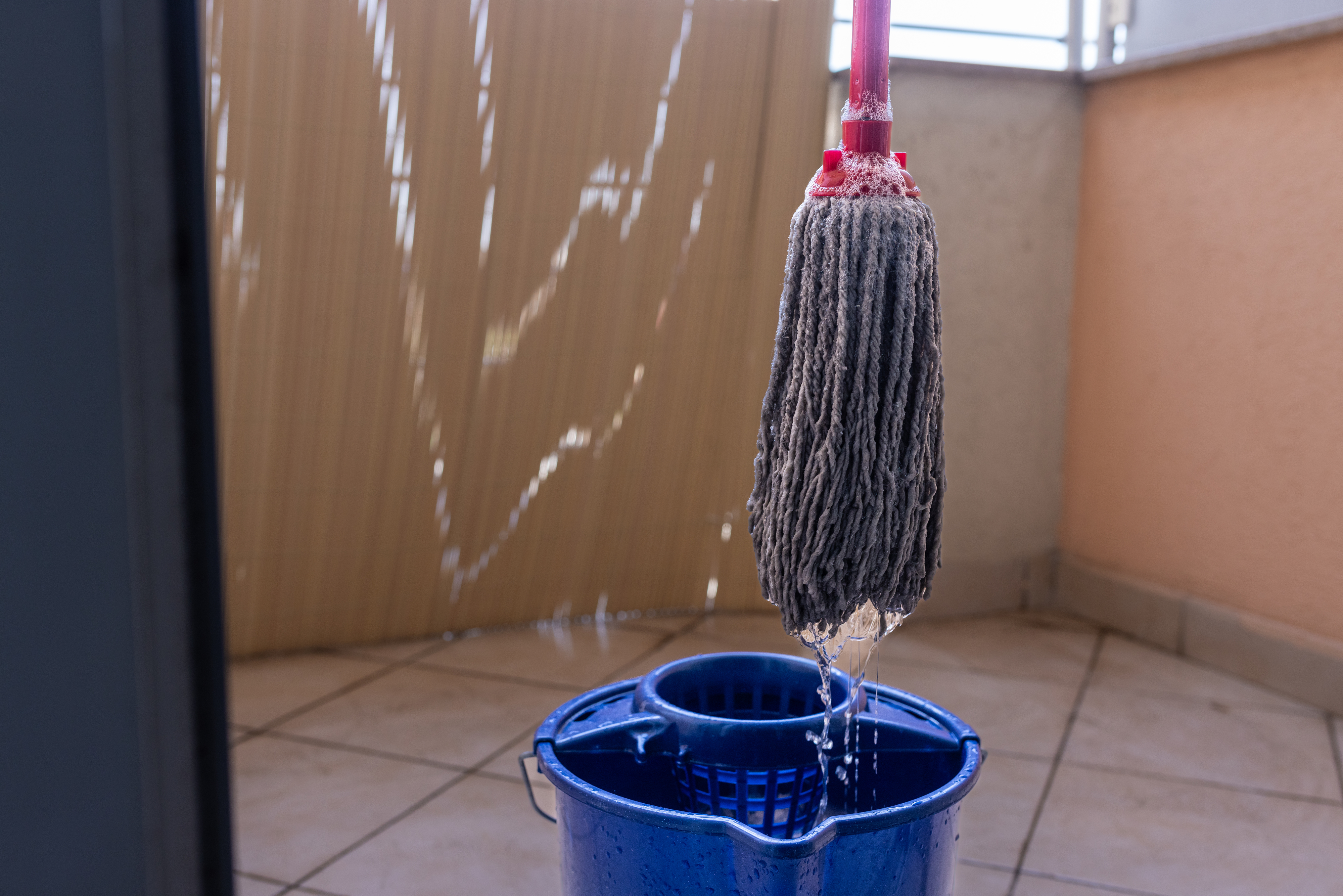 A mop is hanging over a blue bucket, with water dripping from the mop unto the tiled floor below