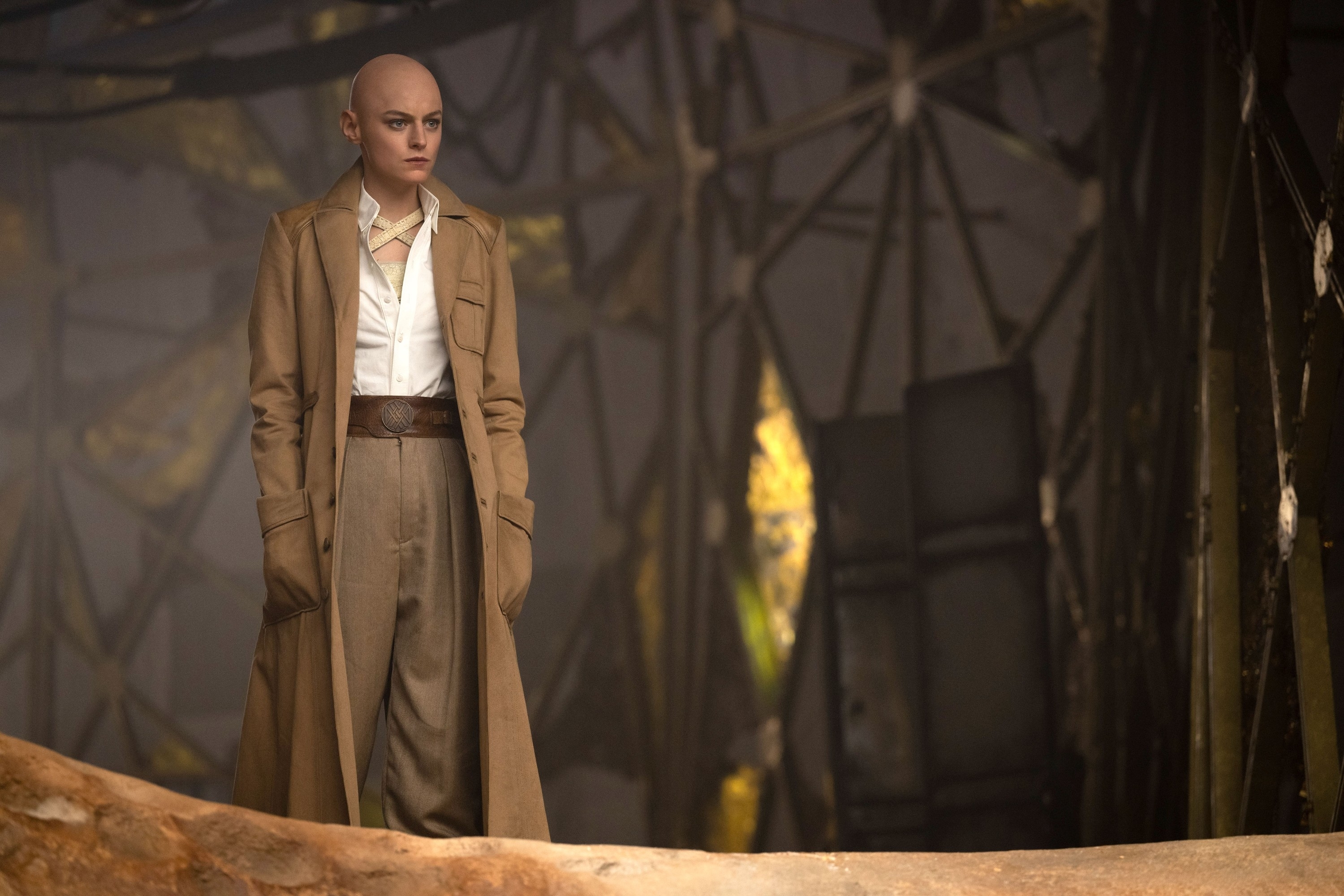 Tilda Swinton, dressed in a long beige coat and loose pants, stands in a mysterious setting with intricate metal frameworks and lighting effects