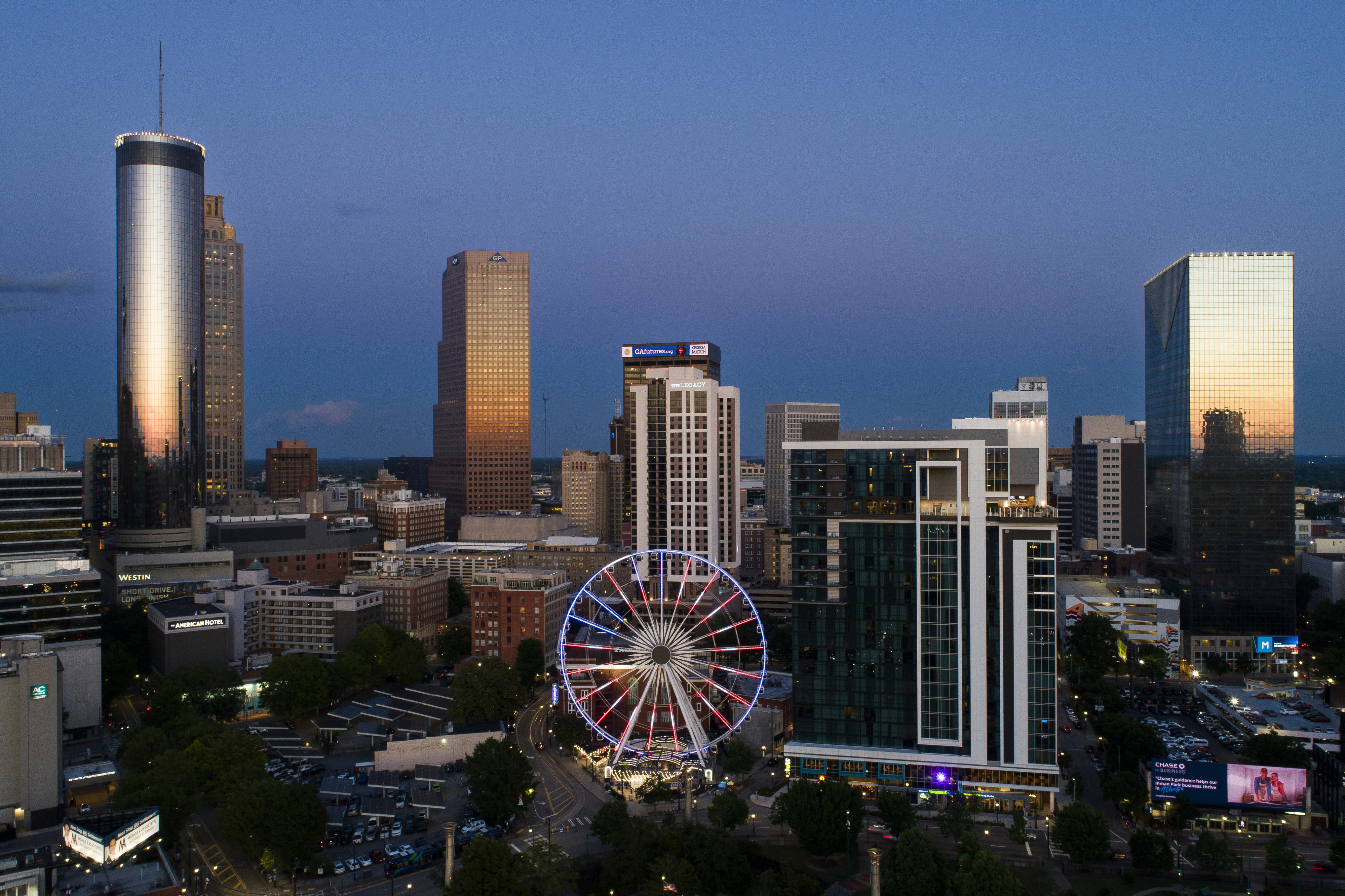 Skyline of a city at dusk featuring tall buildings and a large Ferris wheel in the center. The sky is clear and transitioning to night