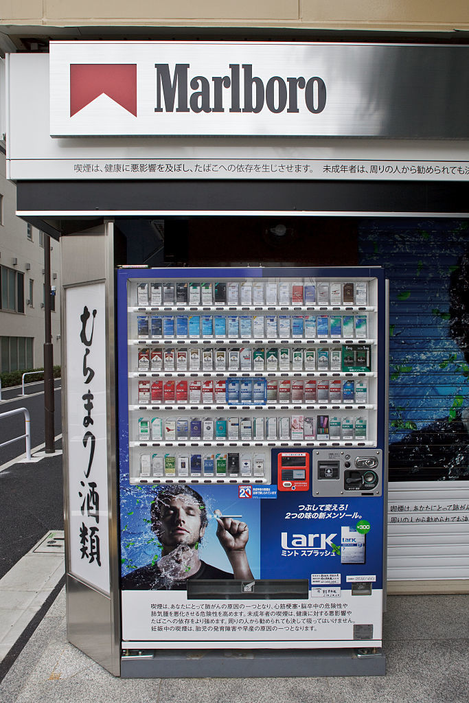 Cigarette vending machine under a Marlboro , displaying various cigarette brands including Lark. Japanese text is on the machine and the sign next to it