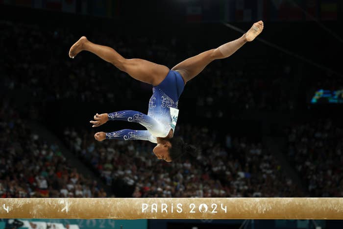 Simone Biles performs on the balance beam during the 2024 Paris Olympics, executing a backflip in front of a large audience
