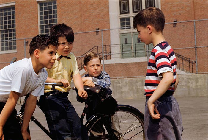 Four boys are talking on a school playground in the 1950s. One boy is sitting on a bike while the other three stand and listen