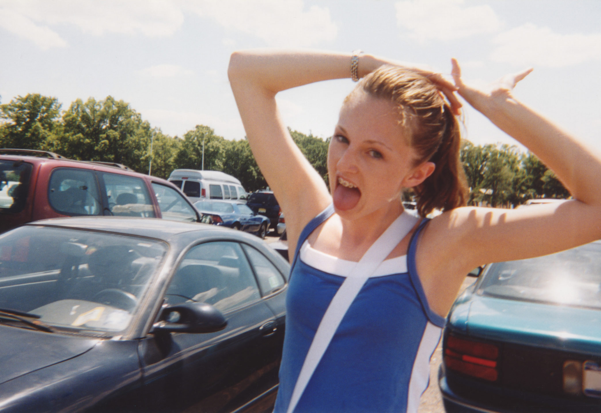 Young woman stands in a parking lot, playfully tugging her ponytail and sticking out her tongue. Numerous cars are visible in the background