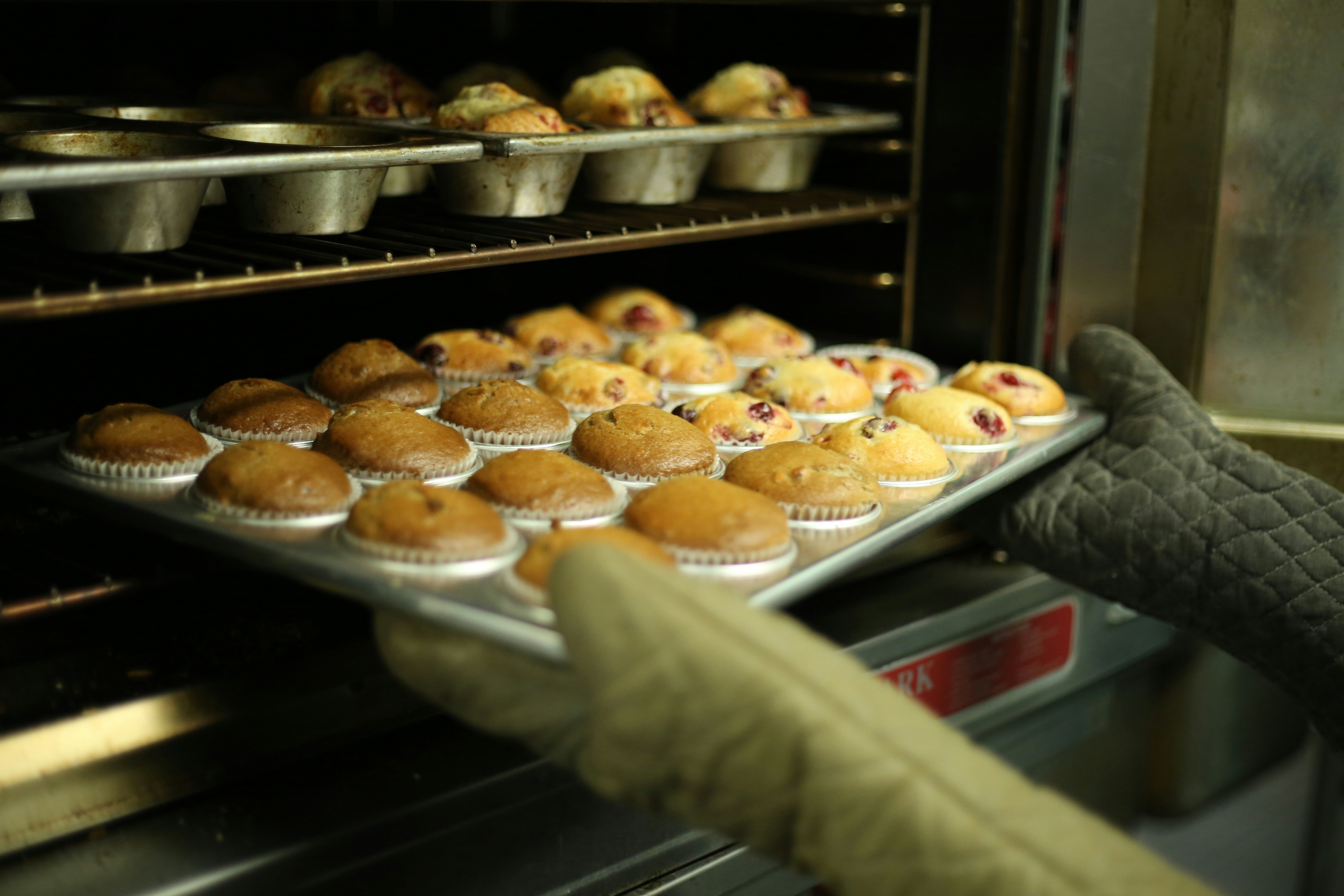 A person wearing oven mitts places a tray of freshly baked muffins, some with visible berries, into an oven with more muffins baking on a rack above