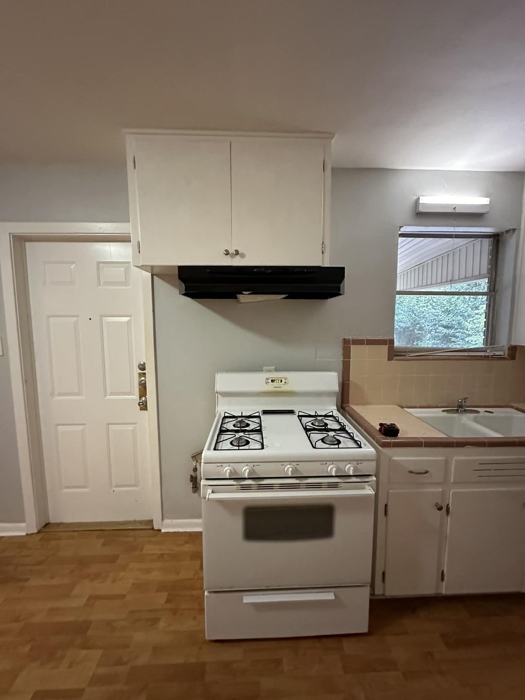 Small kitchen with a white stove, overhead cabinets, and a sink under a window. Wood flooring and back door visible