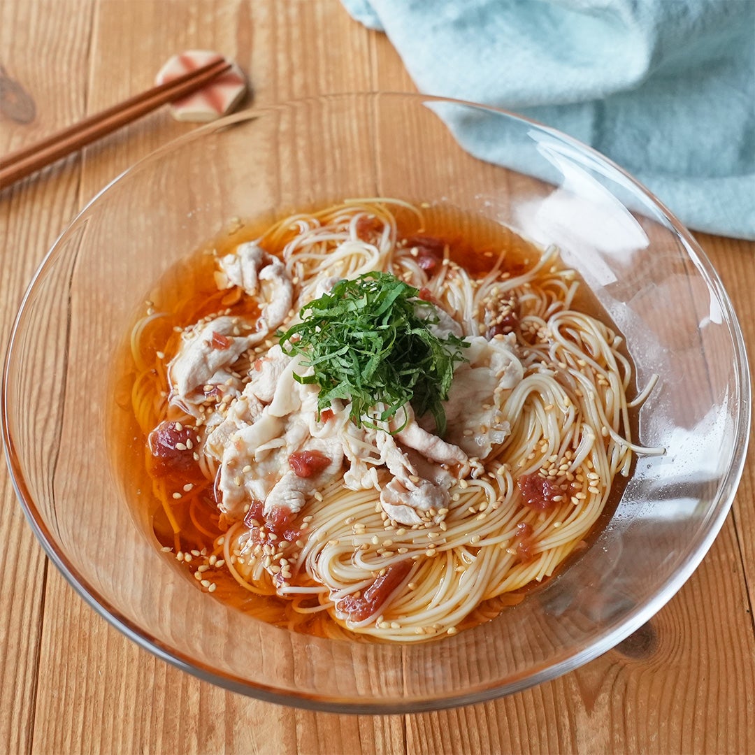 ガラスボウルに入った冷たい素麺の上に豚肉と大葉、胡麻がのった料理。木製のテーブルに置かれ、箸がそばにある。