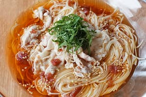 ガラスボウルに入った冷たい素麺の上に豚肉と大葉、胡麻がのった料理。木製のテーブルに置かれ、箸がそばにある。