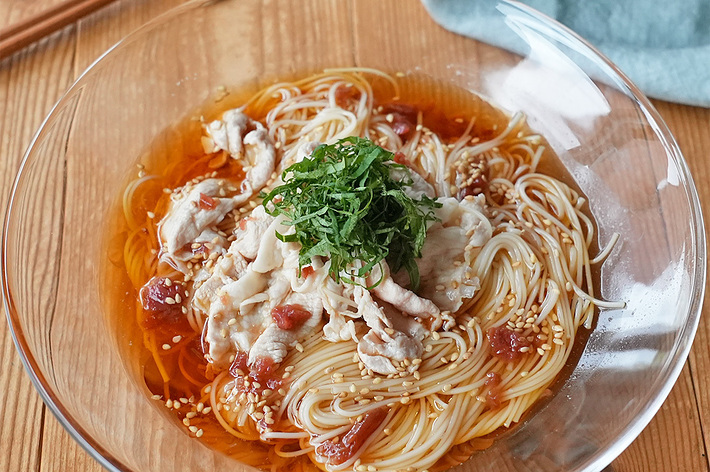 透明なガラスのボウルに盛られた冷麺。上には薄切りの肉、梅干し、大葉、ごまがトッピングされている。木製の箸と布が横に置かれている。