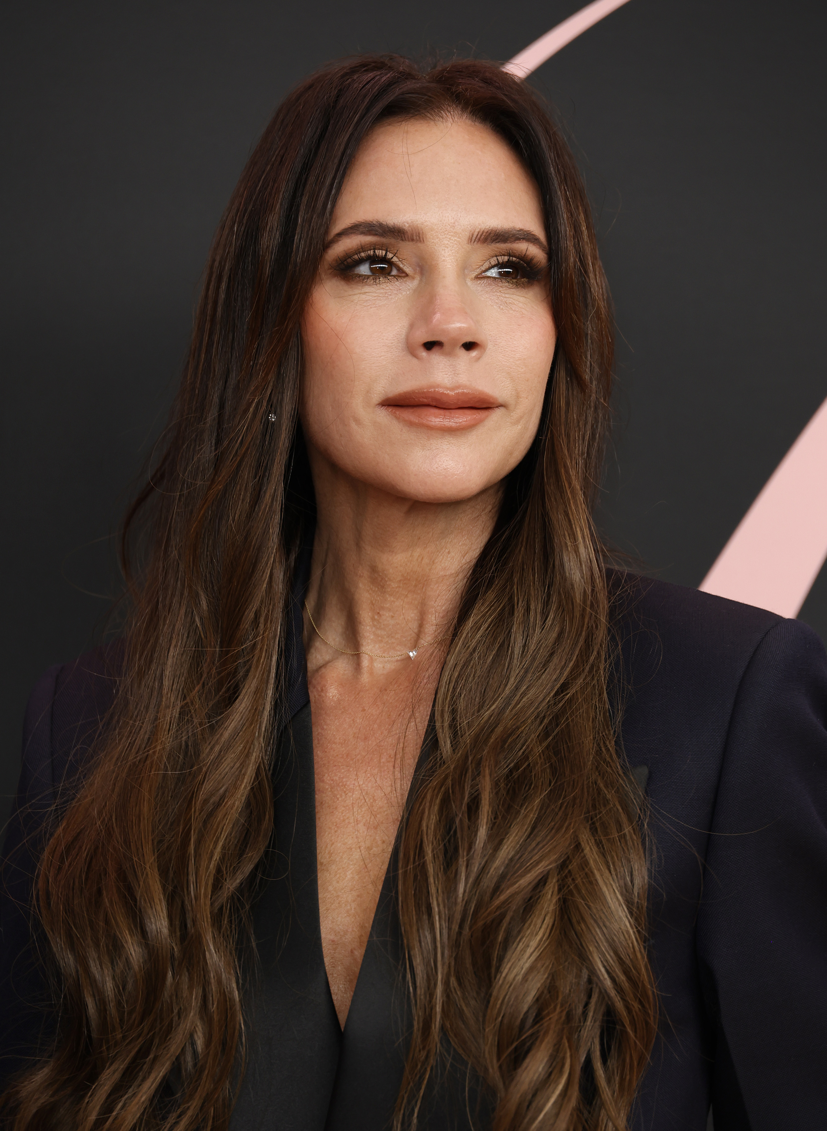 Victoria Beckham with long wavy hair, wearing a black blazer, poses against a solid backdrop