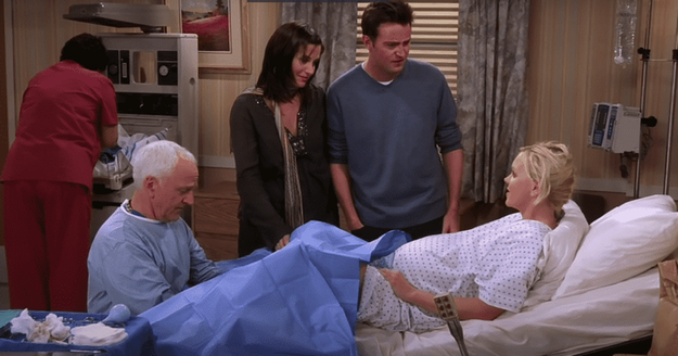 Courteney Cox, Matthew Perry, Lisa Kudrow, and an unidentified actor in a hospital scene from the TV show Friends. Cox and Perry stand beside Kudrow, who is in a hospital bed