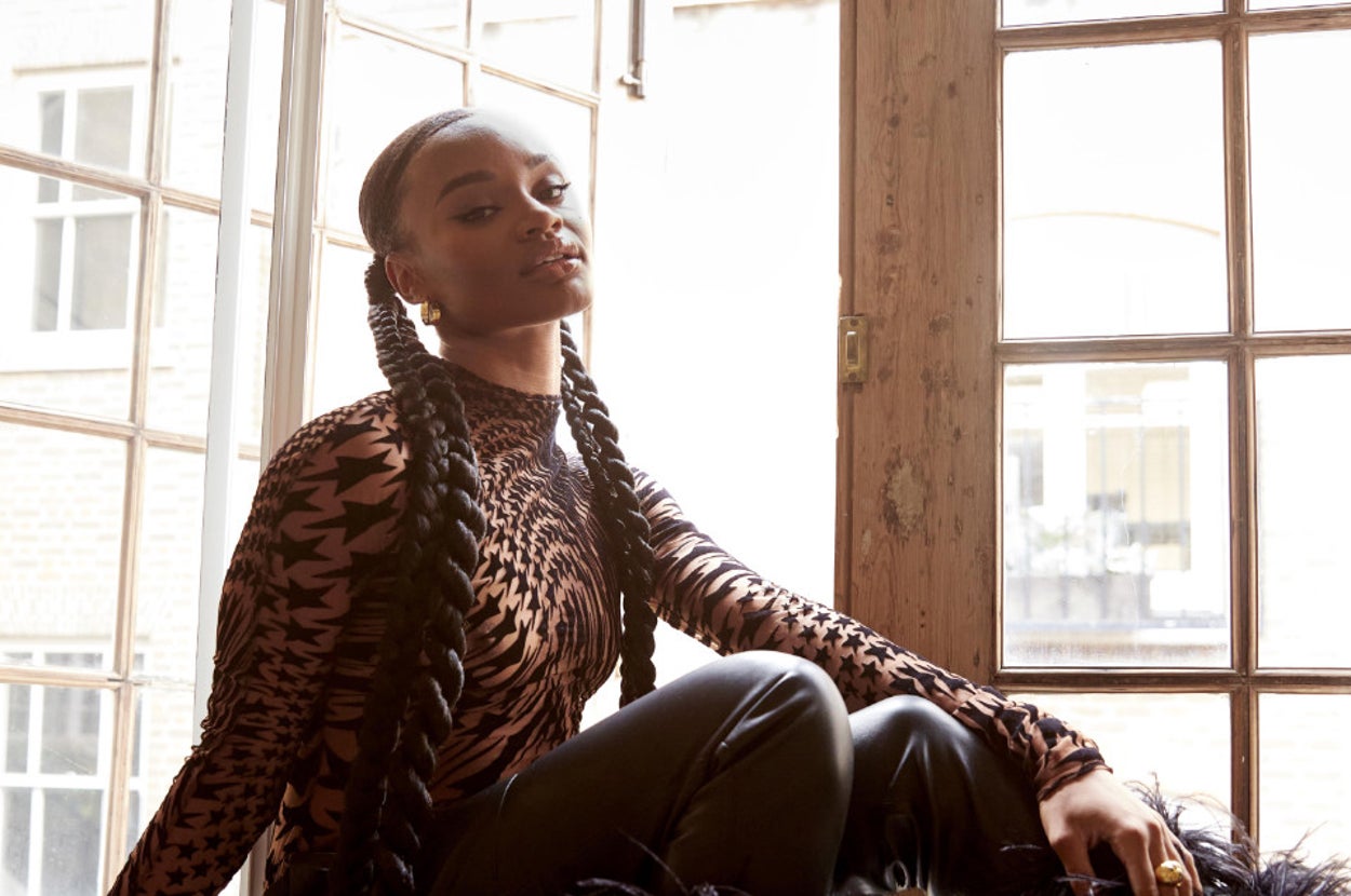 Yara Shahidi sitting by a window wearing a patterned long-sleeve top and black pants with feathered cuffs