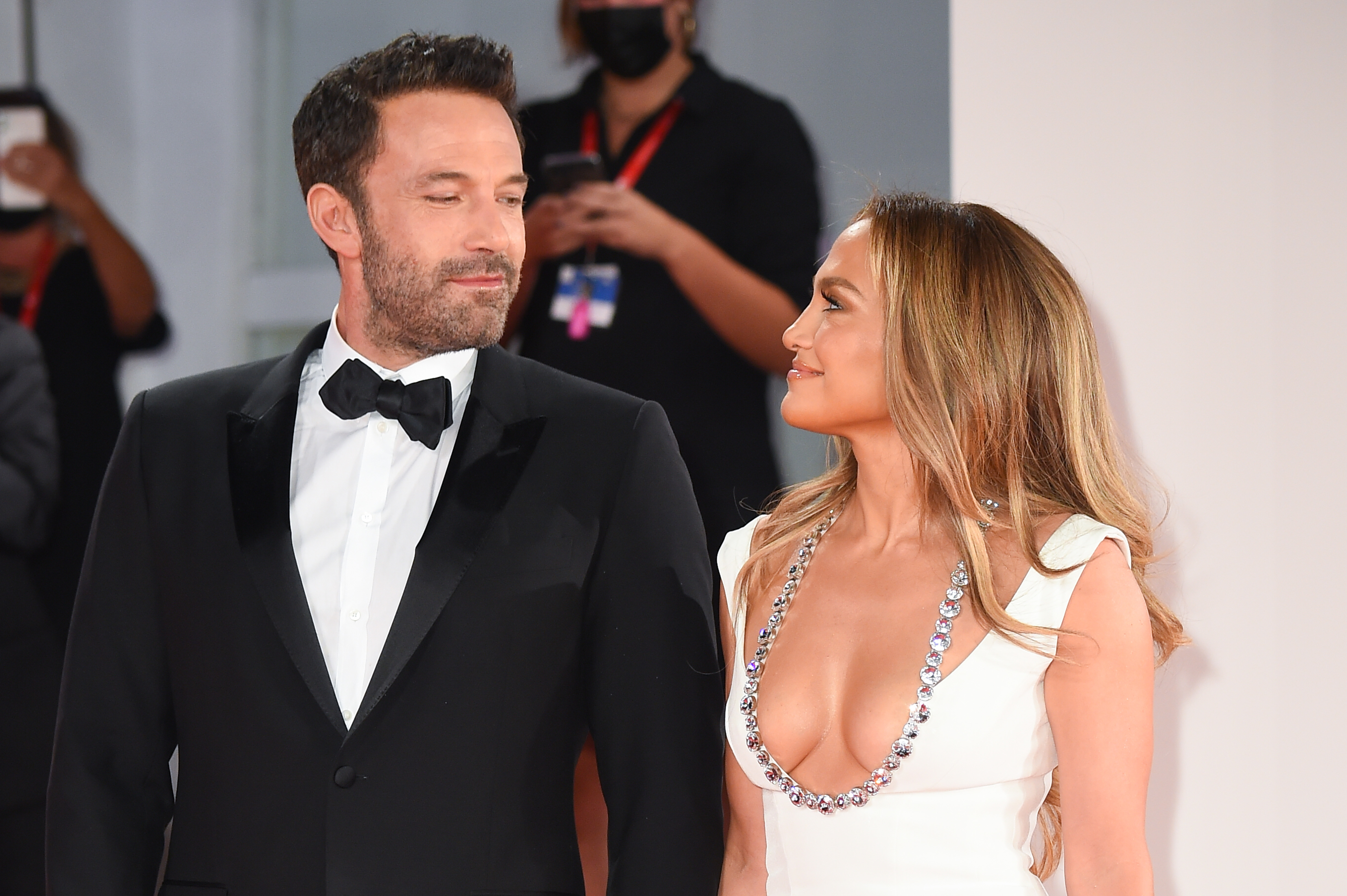 Ben Affleck and Jennifer Lopez at a formal event. Ben wears a black tuxedo with a bow tie, Jennifer wears a plunging white gown with silver embellishments