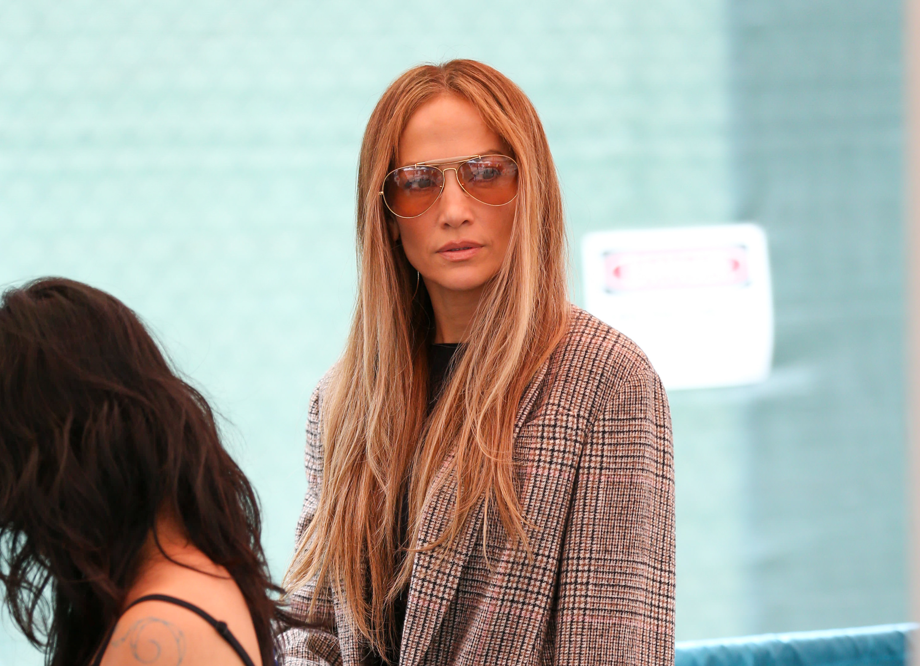 Jennifer Lopez wearing a plaid jacket and sunglasses, standing next to a seated person