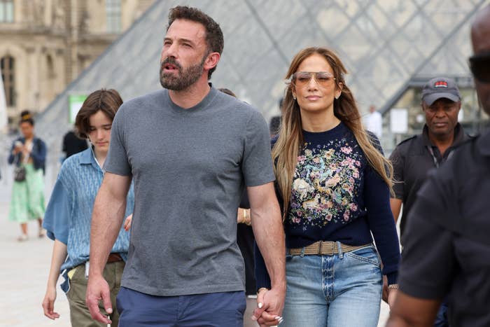 Ben Affleck and Jennifer Lopez walk hand in hand outdoors. Jennifer is in jeans and a floral top, while Ben wears a gray T-shirt and blue pants