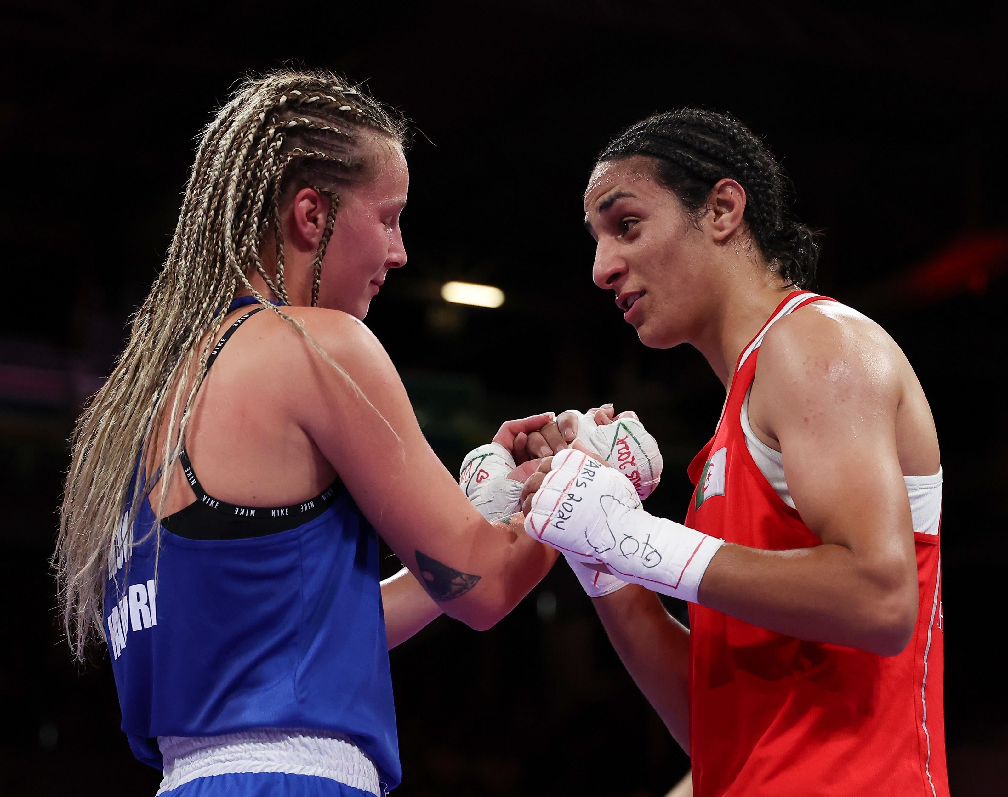 Boxers Imane Khelif and Amy Broadhurst tap gloves in a congratulatory gesture after a match in an arena. Khelif in red, Broadhurst in blue