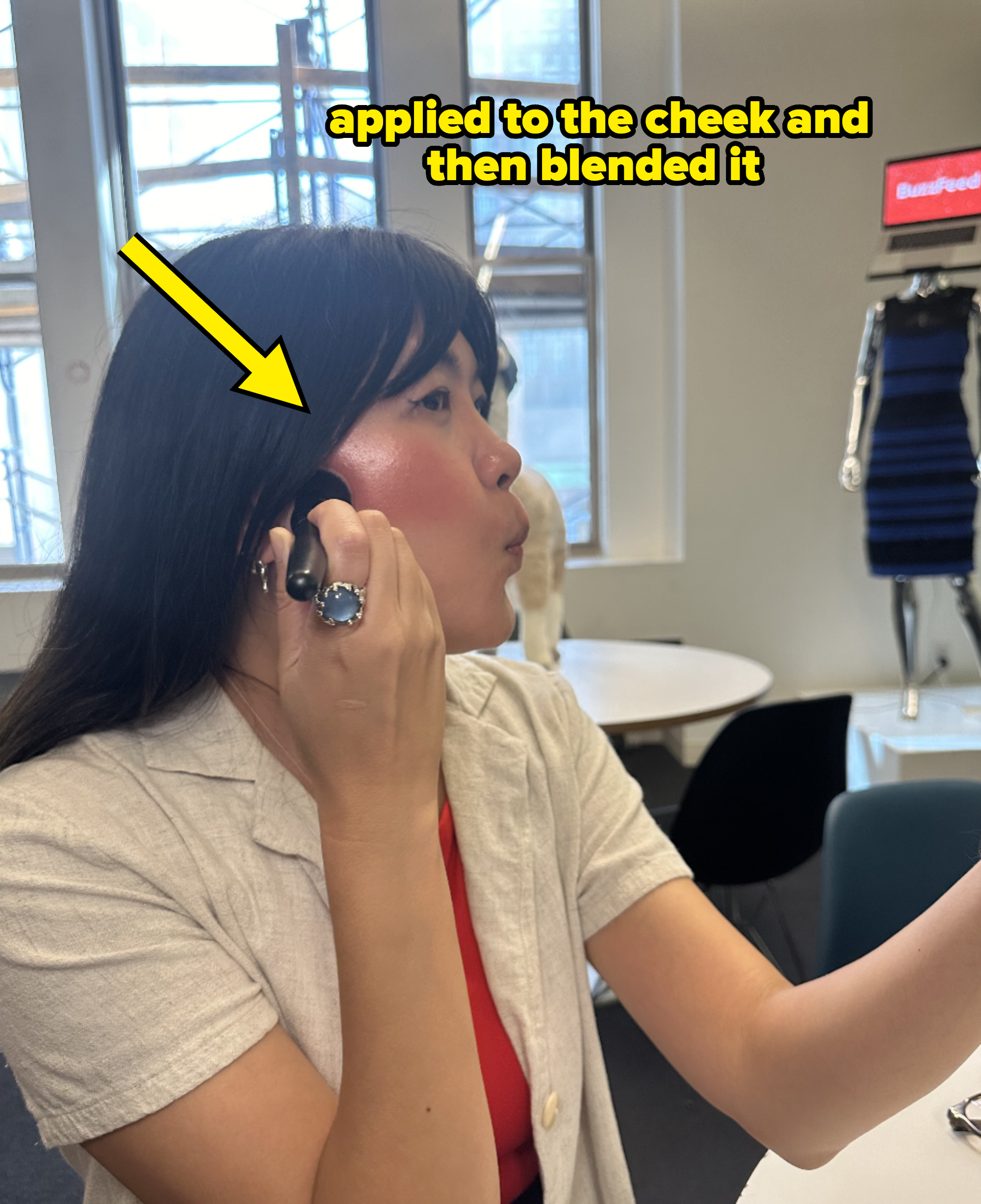 Woman applies makeup in an office with a mannequin and a "BuzzFeed" sign in the background