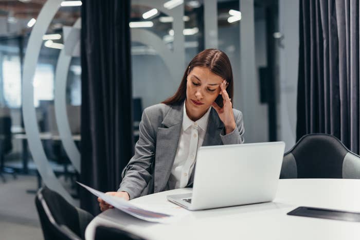 A woman in a grey blazer and white shirt sits at a table in an office, looking stressed while holding a document and sitting in front of a laptop