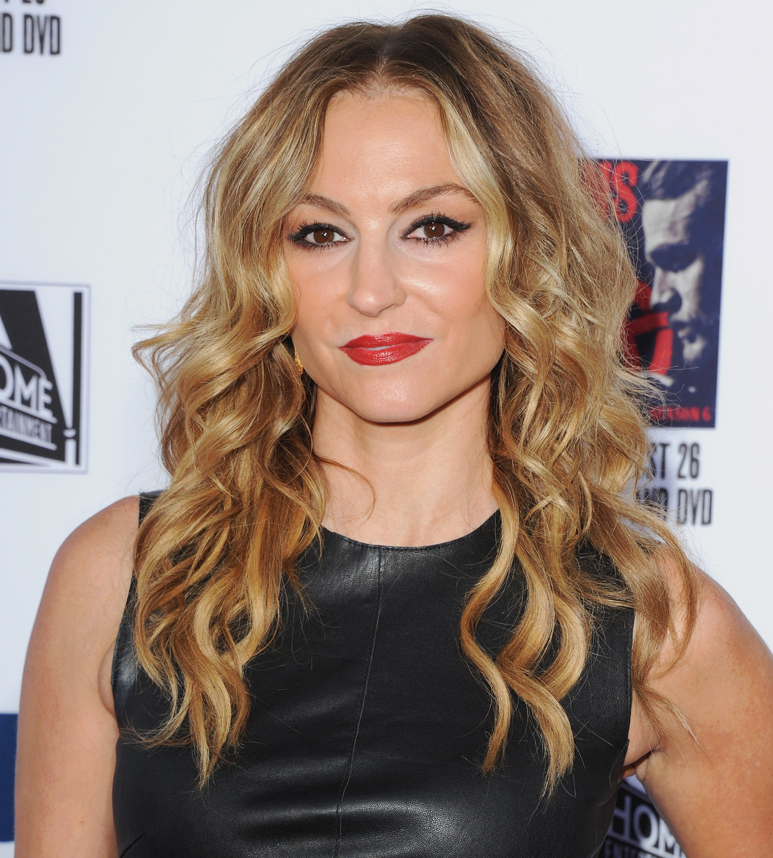 A woman with long, wavy blonde hair wearing a sleeveless black leather top poses for a photo at an event