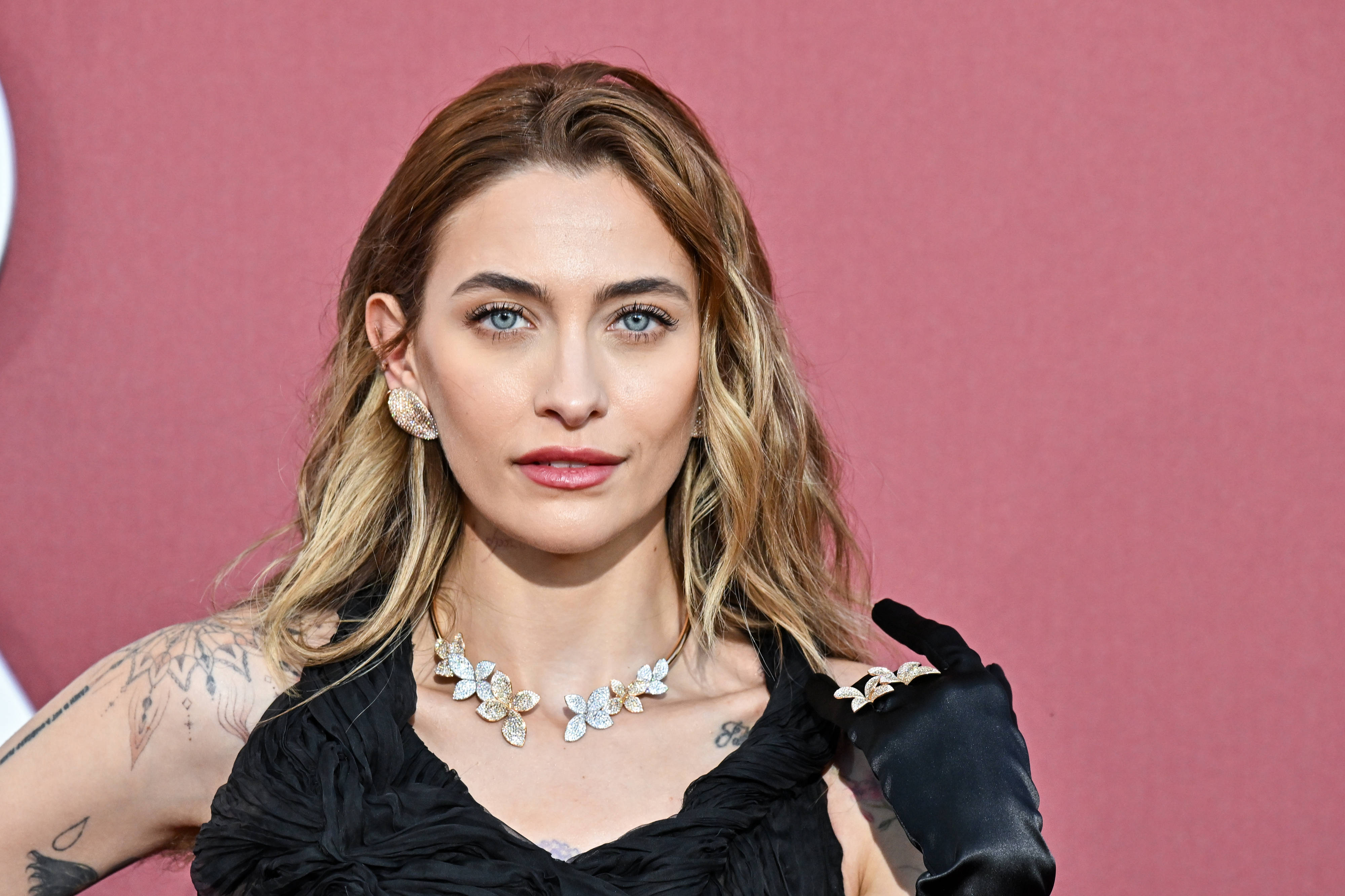 Paris Jackson posing on the red carpet in a black dress with ruffles and gloves, wearing a floral necklace and large earrings