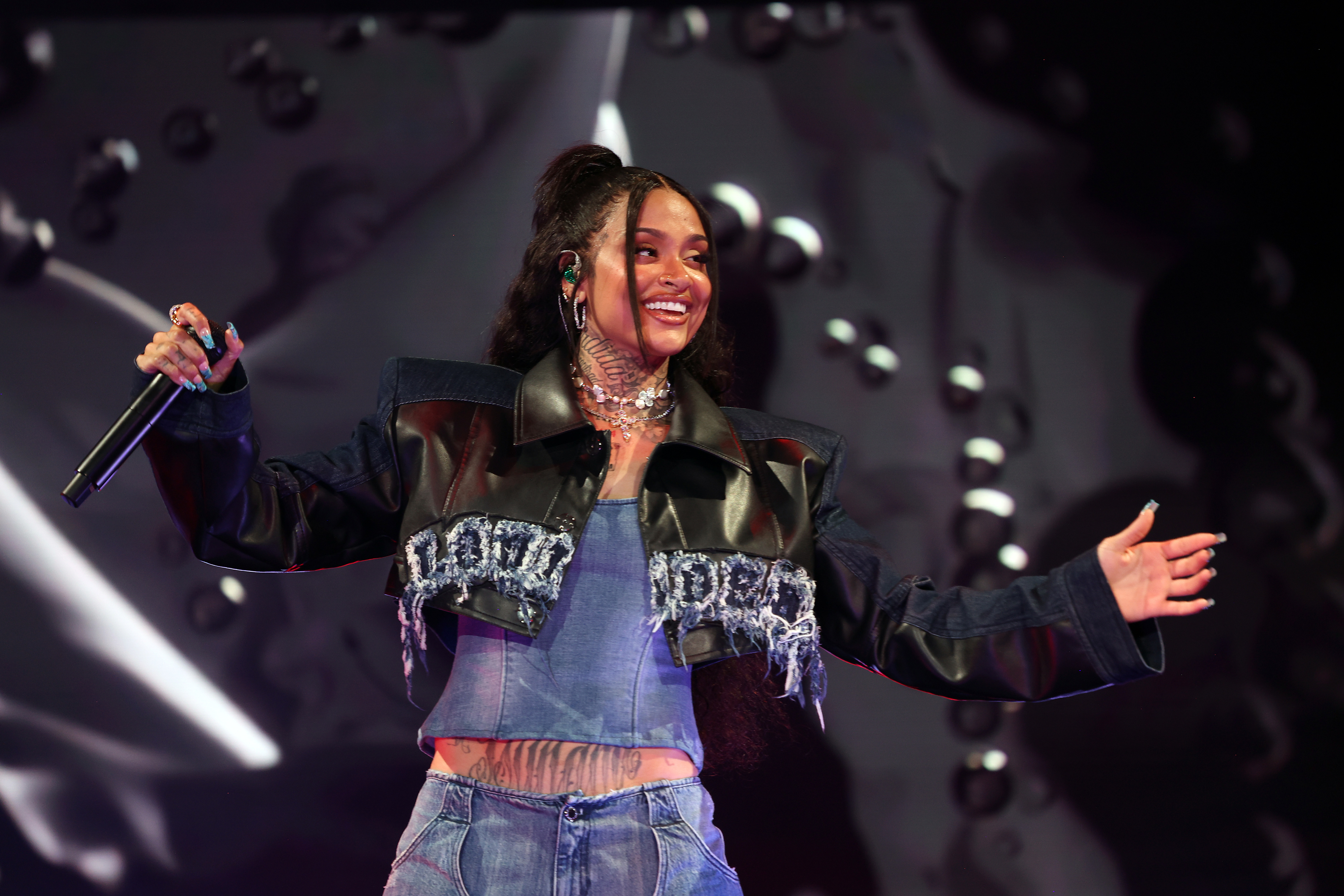 Kehlani performs on stage wearing a denim outfit with a black leather jacket adorned with jeweled embellishments, holding a microphone and smiling