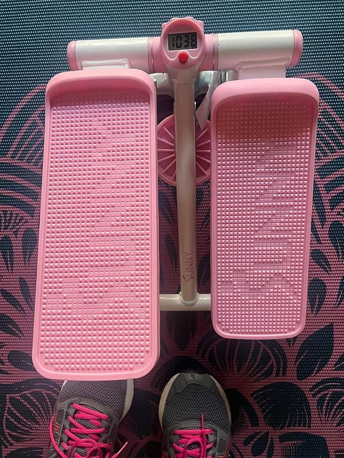 Image of a pink step machine with a digital display showing 10:38. Below, a person in sneakers with pink laces is standing on a patterned mat