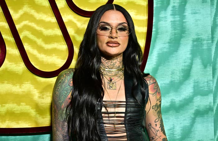 Kehlani poses in a black, strappy outfit with long flowing hair and visible tattoos on a patterned background