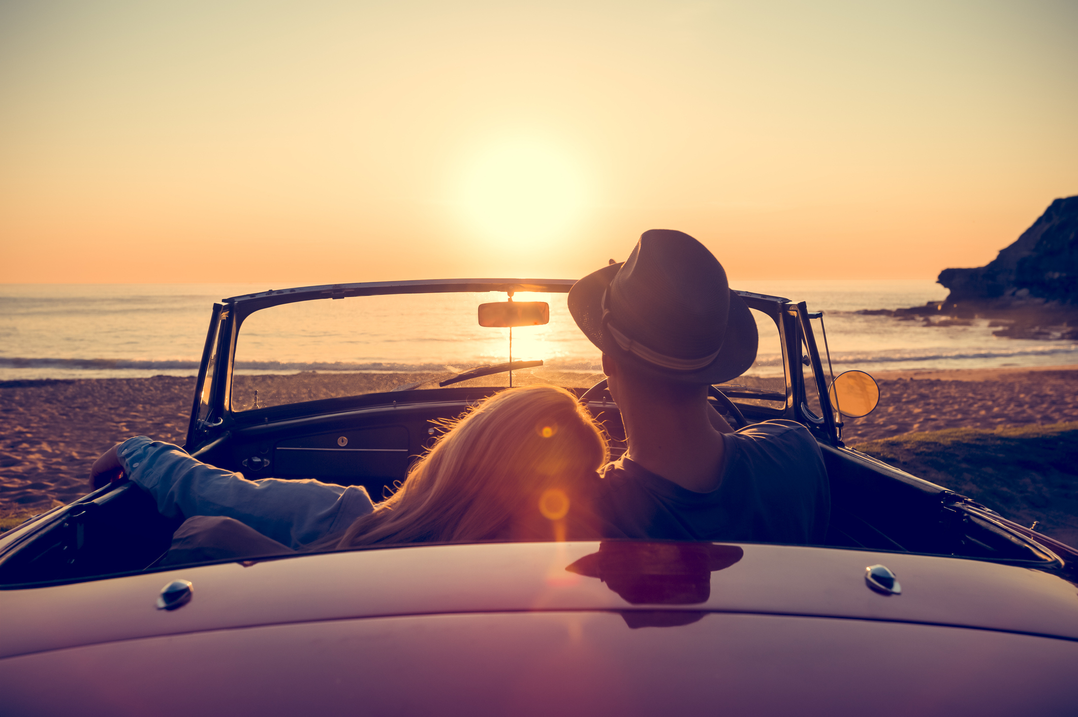 A couple is sitting in a convertible car at the beach during sunset, embracing each other affectionately and gazing at the horizon