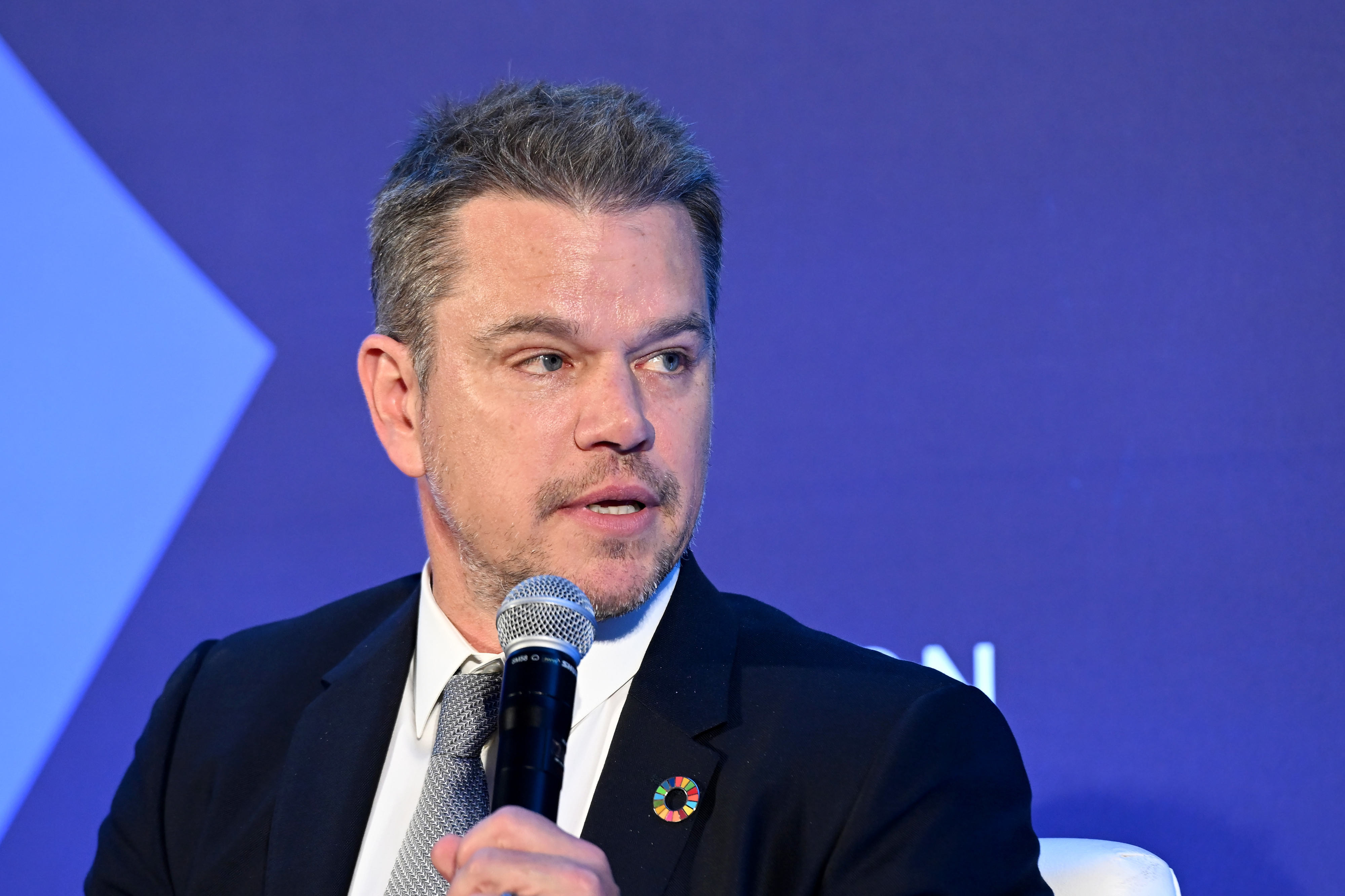 Matt Damon is speaking into a microphone at a formal event, dressed in a suit and tie