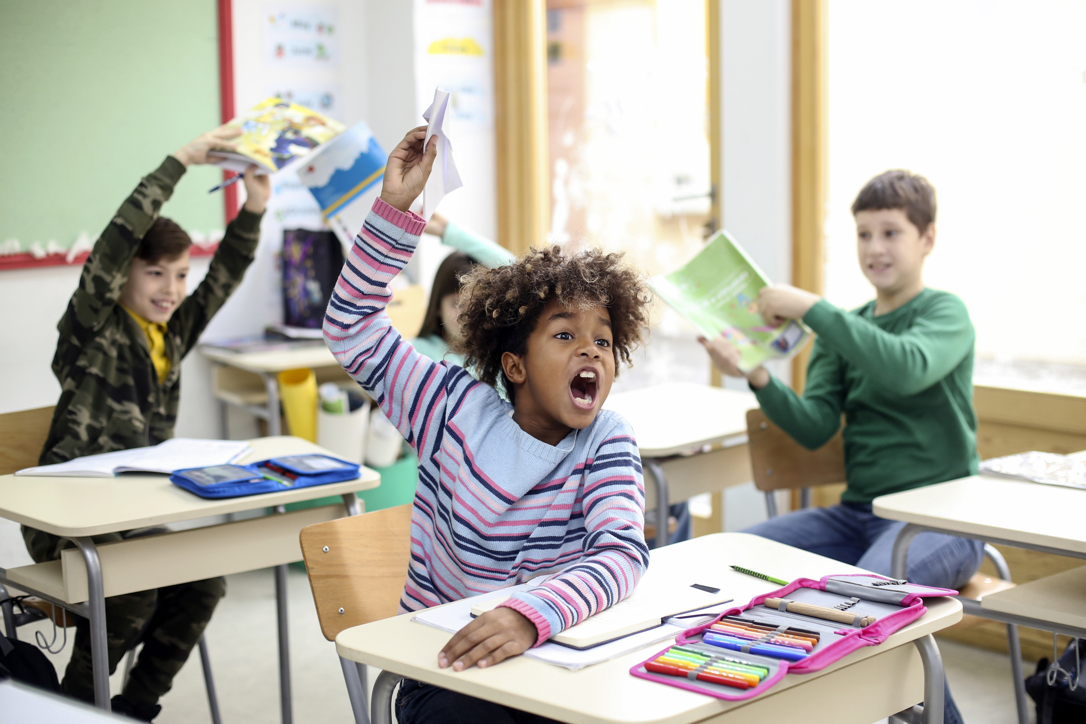 Children in a lively classroom; one child in the foreground enthusiastically raises their hand, while others hold up books and papers