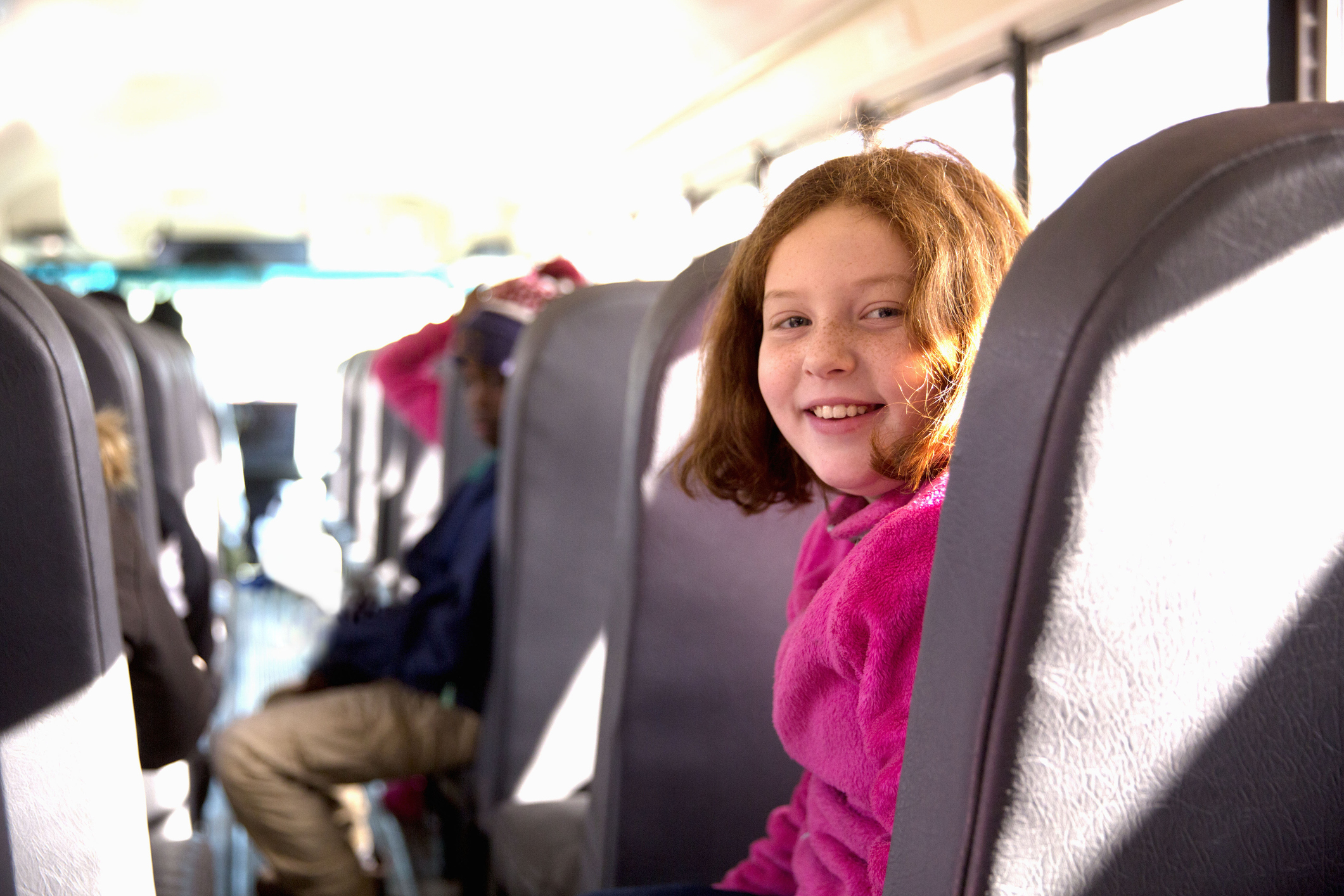 A young girl with red hair and a bright pink sweater smiles while sitting in a school bus seat. Other children are visible in the background