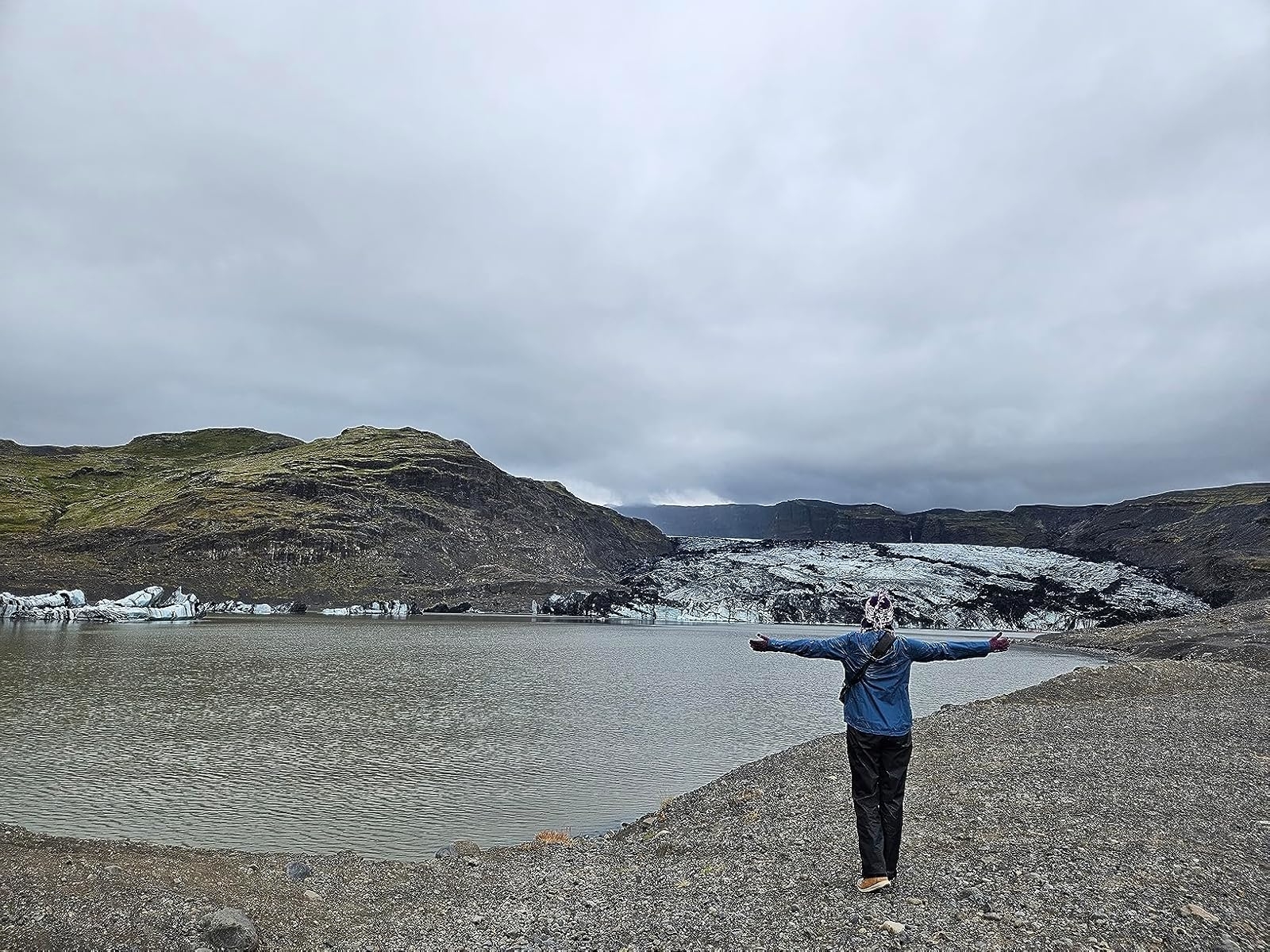 A person stands with arms outstretched facing a glacial lagoon, with rocky terrain and mountains in the background, wearing the blue raincoat