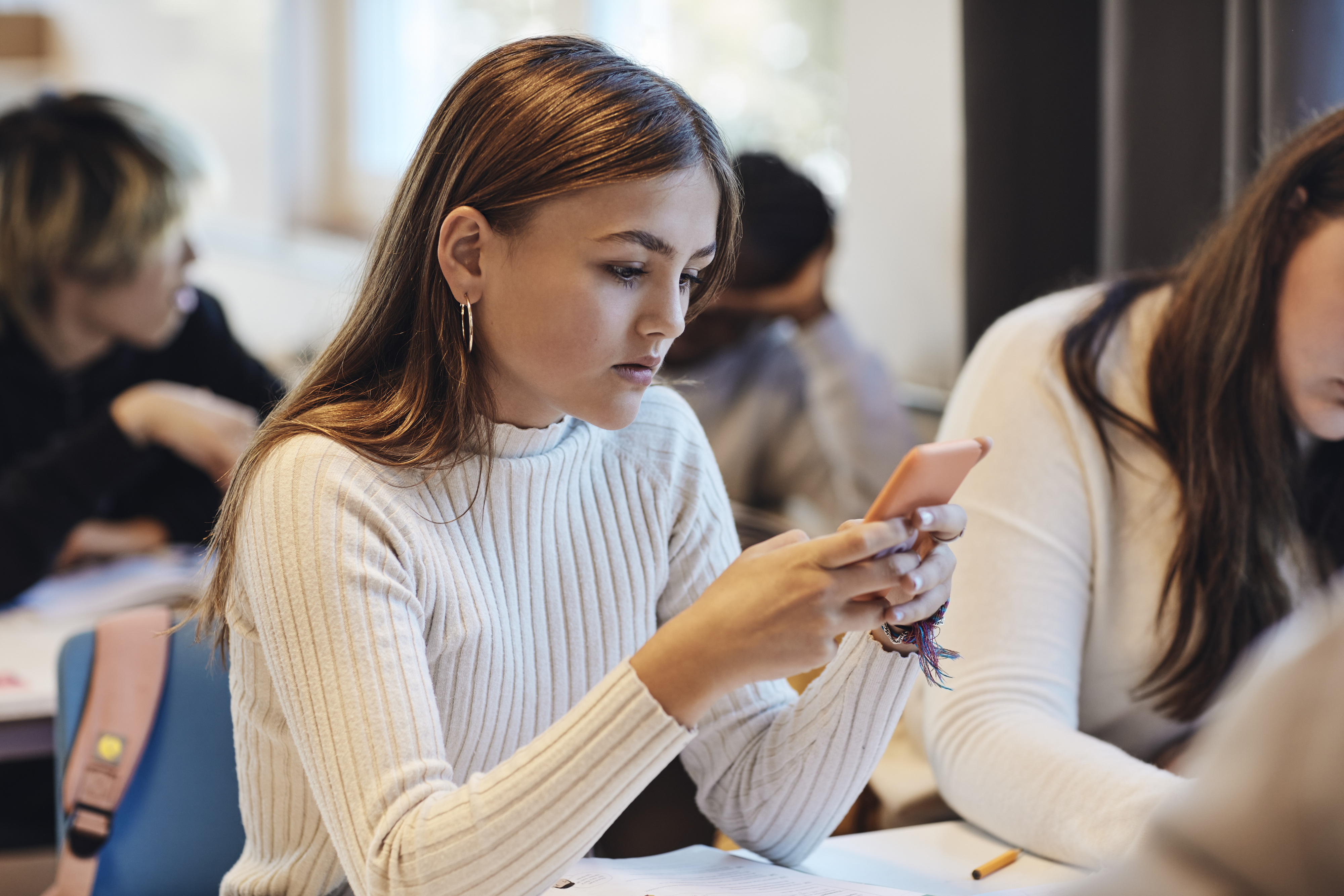 A young woman is focused on her phone in a classroom setting, with other students in the background. Names of the individuals are not known