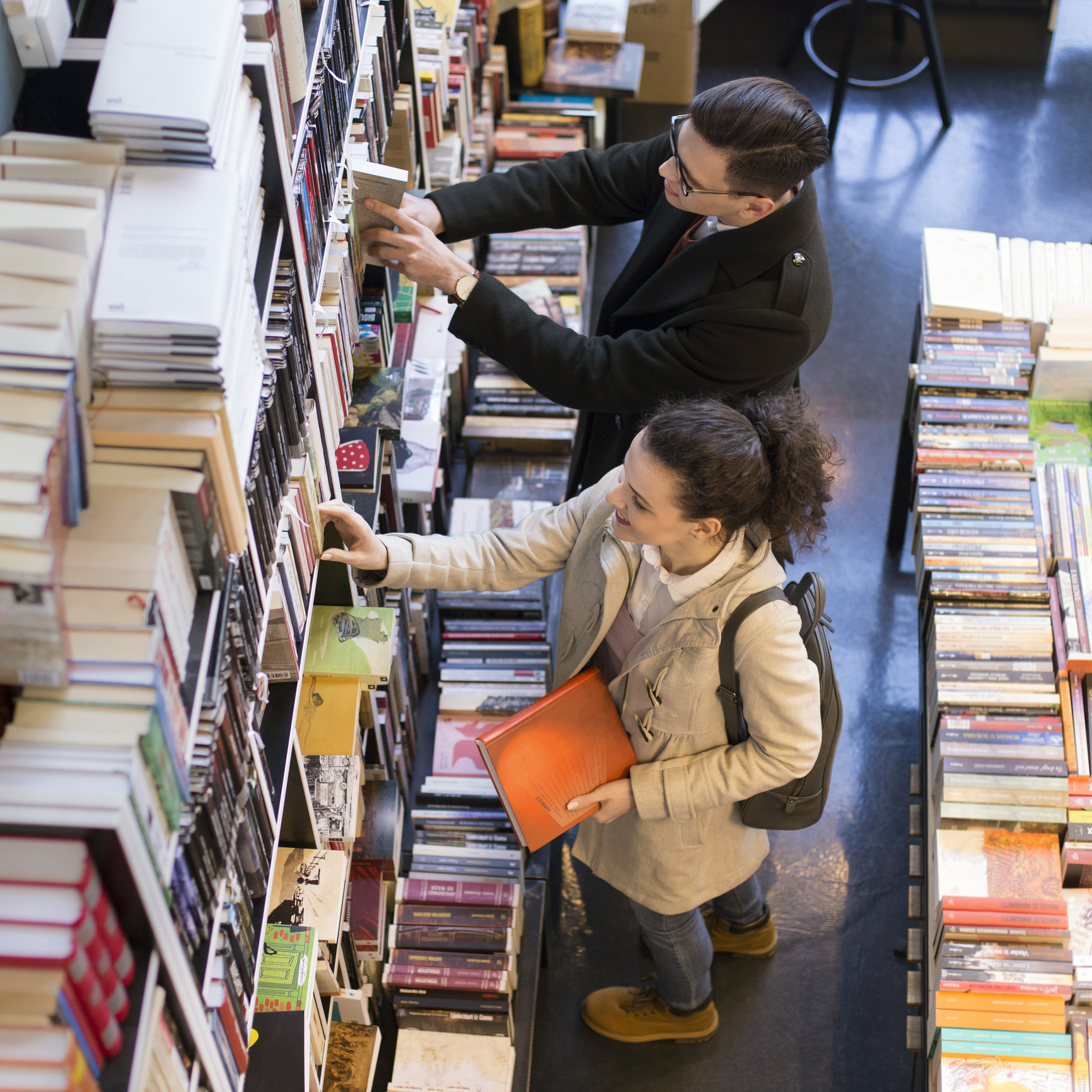 A man and woman browse a bookstore, picking books from shelves. The woman holds an orange book and has a backpack. The man wears a coat and glasses
