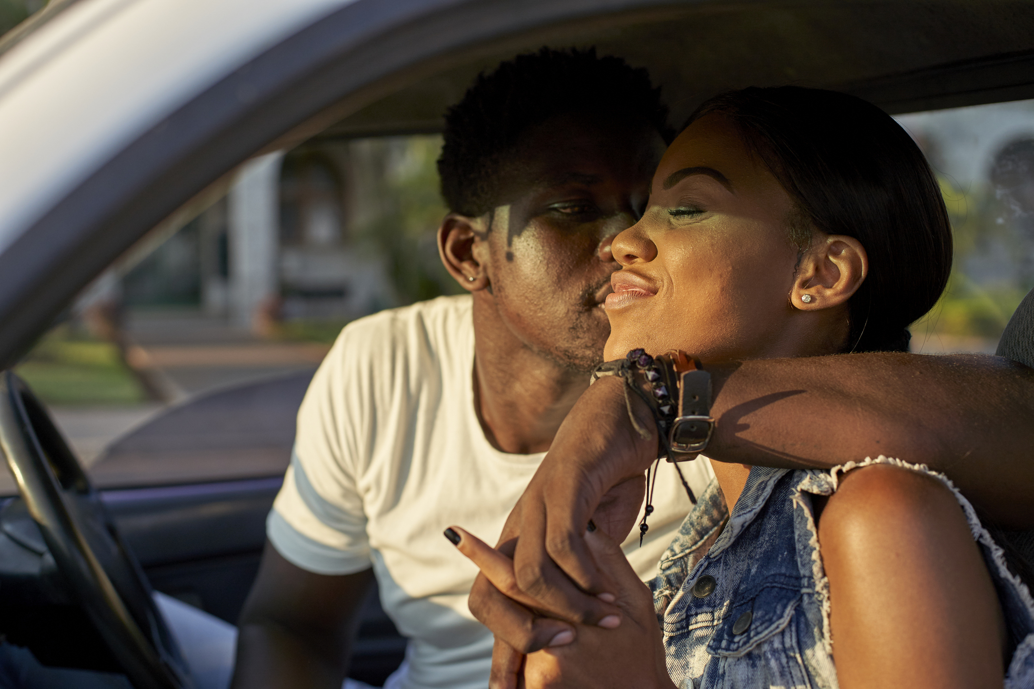 A couple sits in a car, with the man leaning in to kiss the woman's cheek. The woman is smiling, and both appear relaxed and affectionate