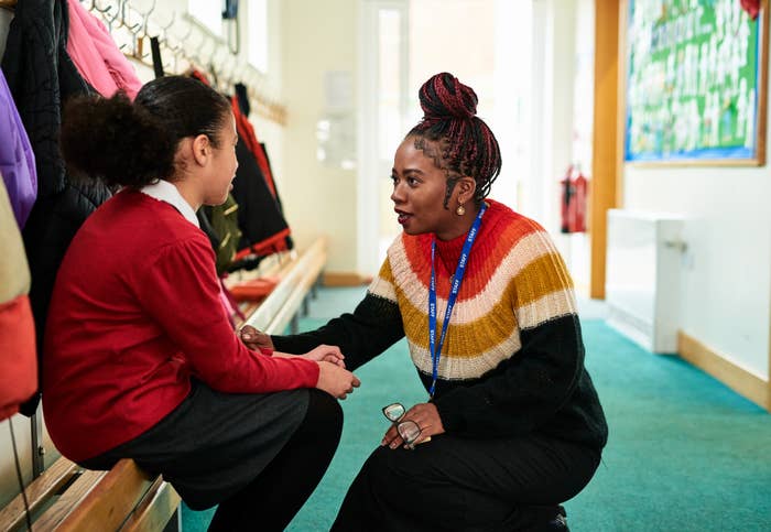 A teacher kneels and talks to a young student in a school hallway, holding the student's hand and offering support
