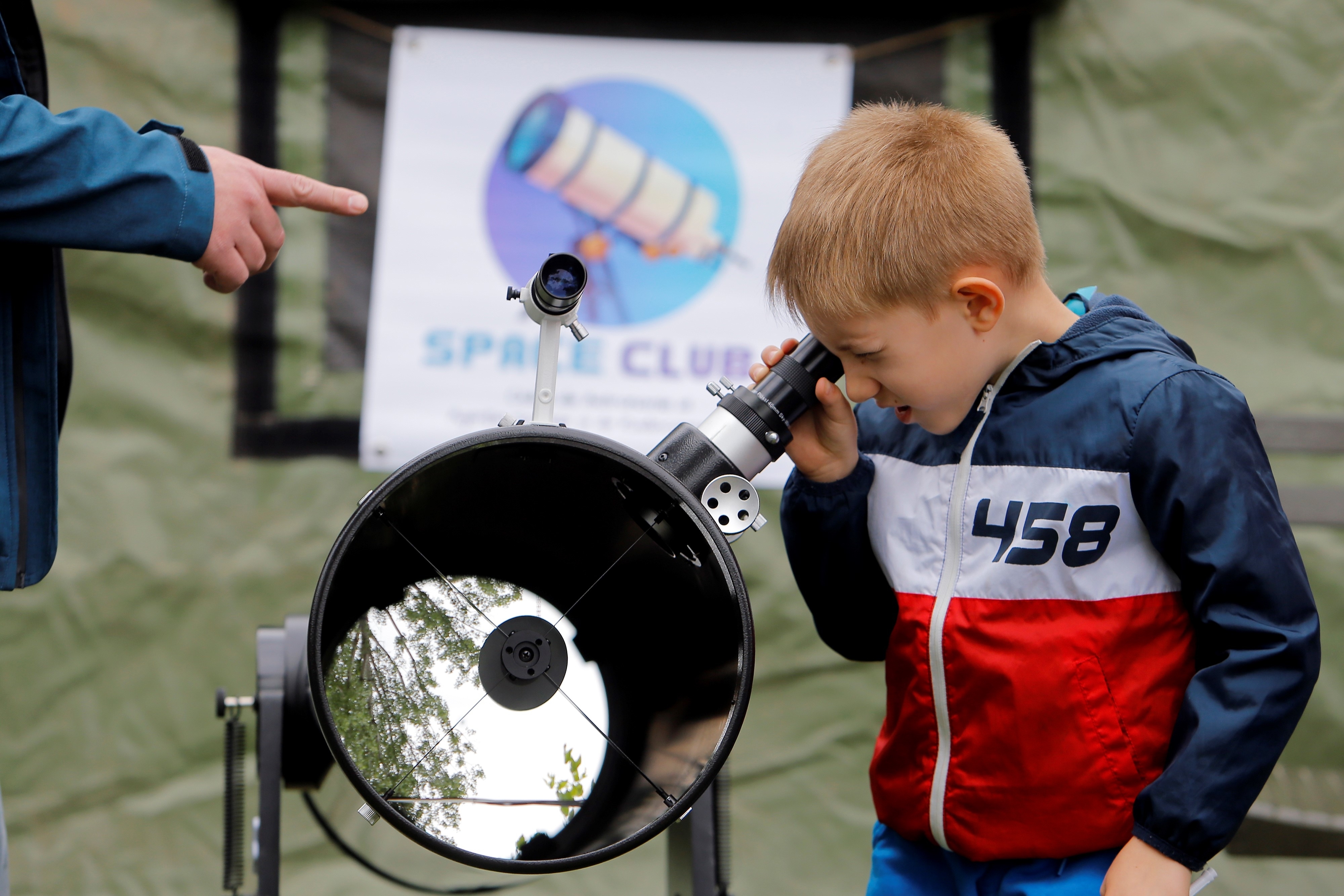 A young boy, wearing a jacket with &quot;458&quot; on it, looks through a telescope at a &quot;Space Club&quot; event, while an adult hand points in the direction he is looking
