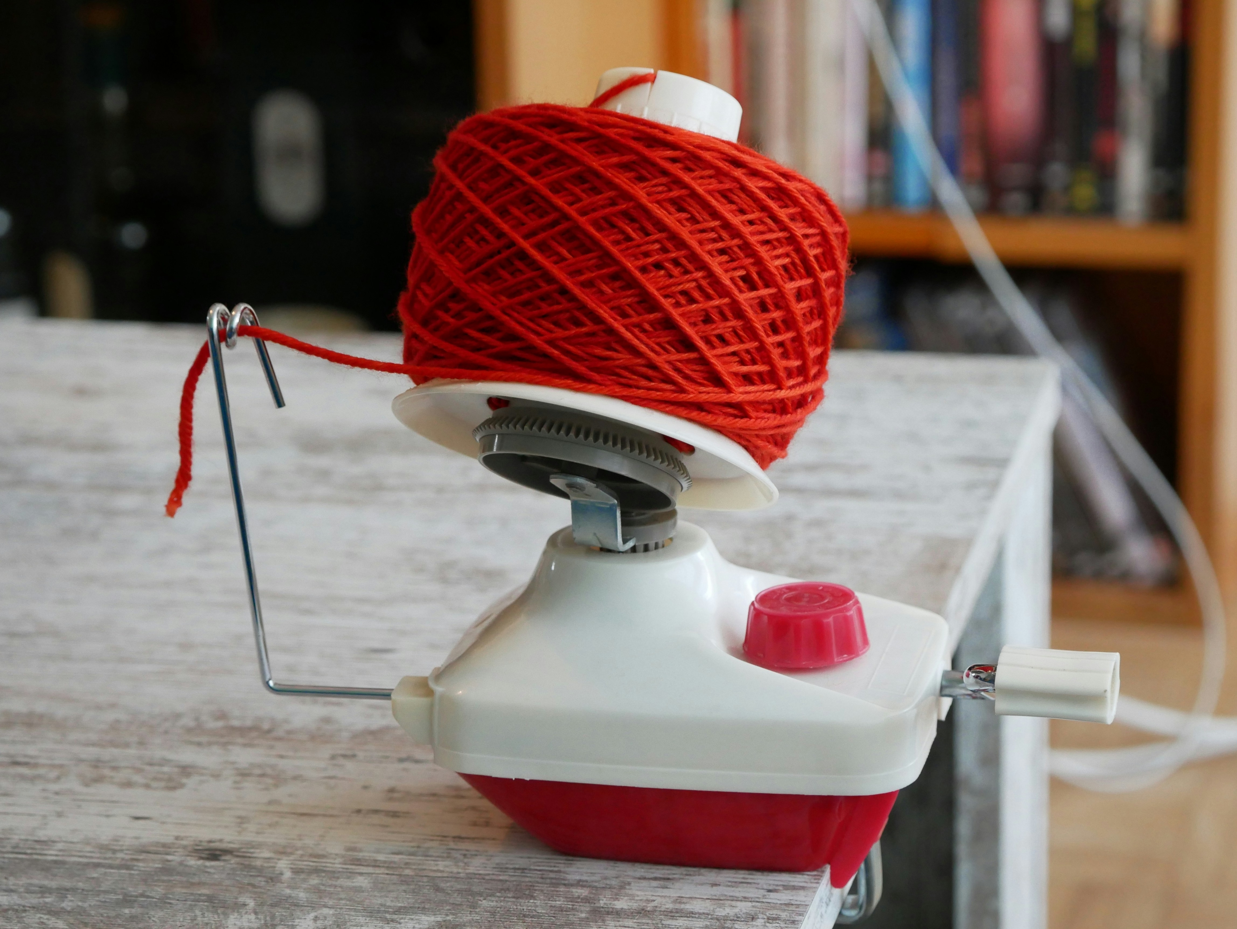 A yarn ball winder secures a red yarn ball on a table. Background includes shelves with books and a visible white cable
