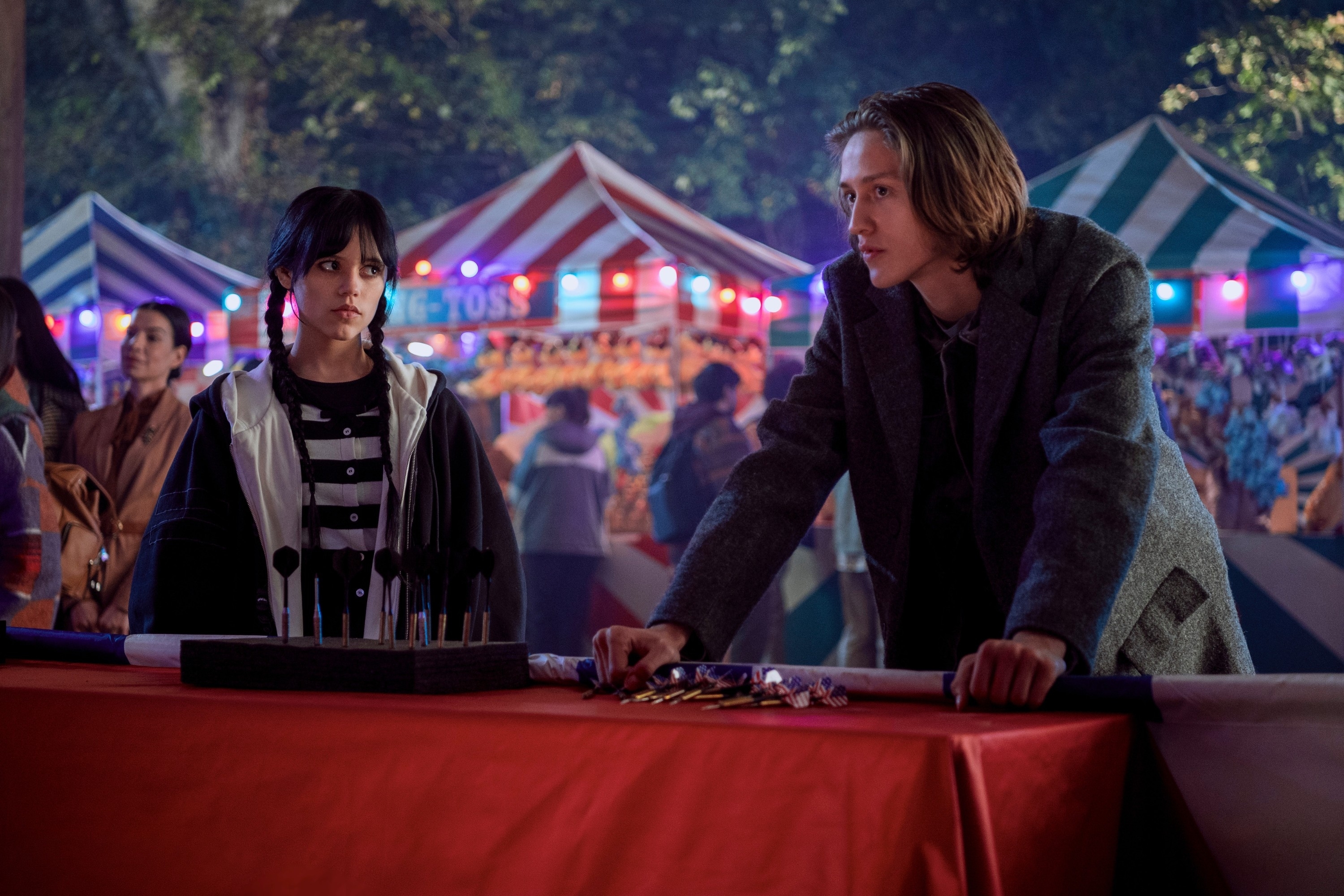 Jenna Ortega and Percy Hynes White in a carnival scene, standing by a game booth with colorful tents and lights in the background