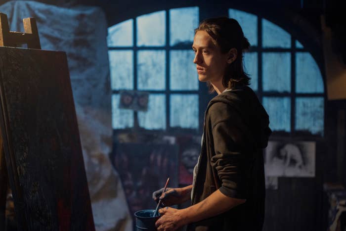 A person stands in an art studio, holding a paintbrush and a container. Various artworks are visible in the background