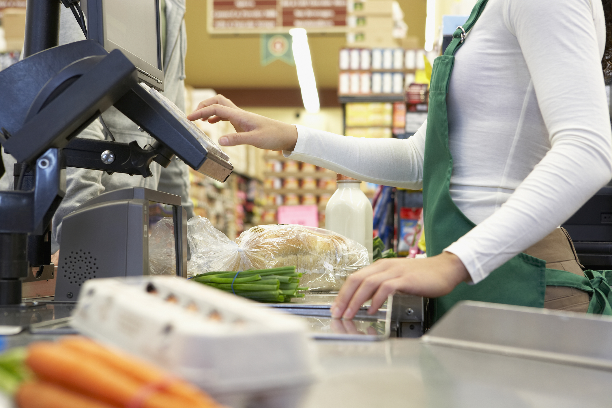 Cashier scanning groceries, including carrots, eggs, green onions, milk, and bread, in a supermarket checkout lane