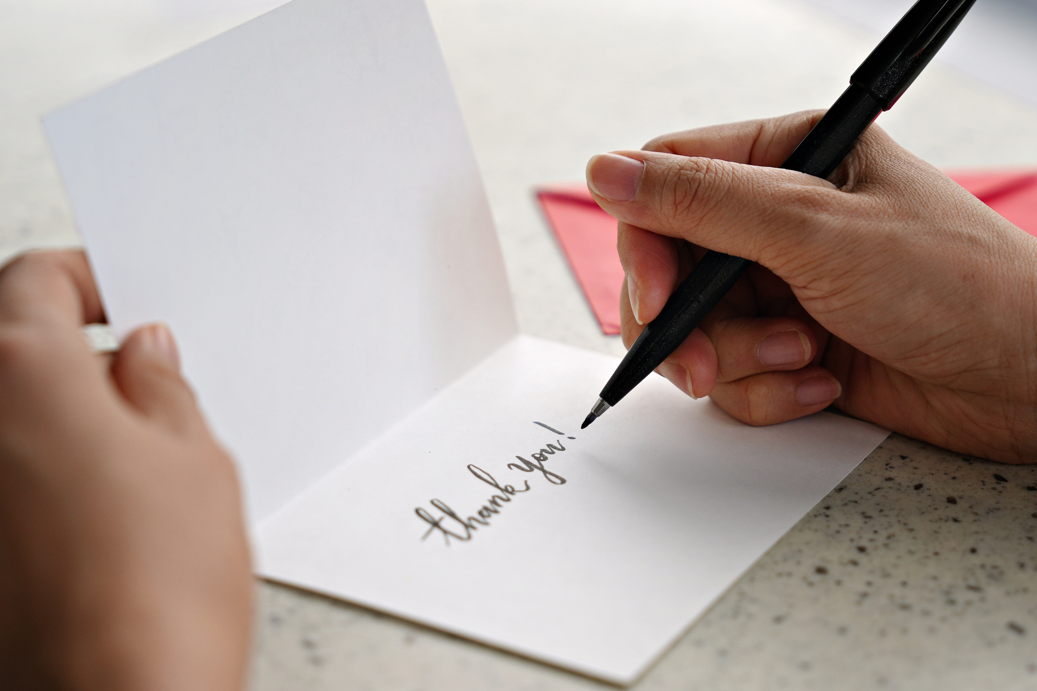 A person writes &quot;thank you!&quot; on a card, holding the card open on a speckled surface