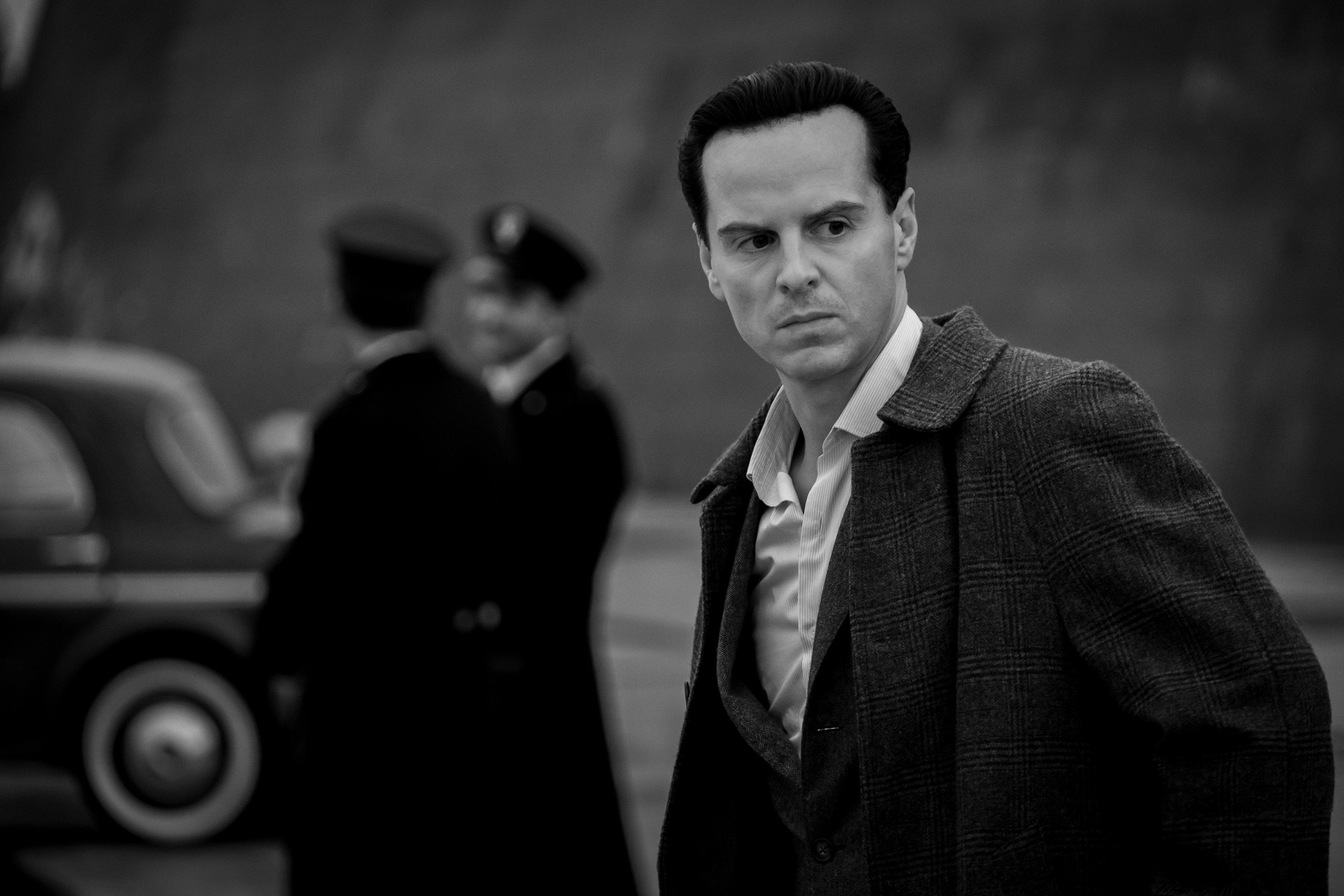 Andrew Scott, in period costume, stands in a serious pose with officers and a vintage car blurred in the background