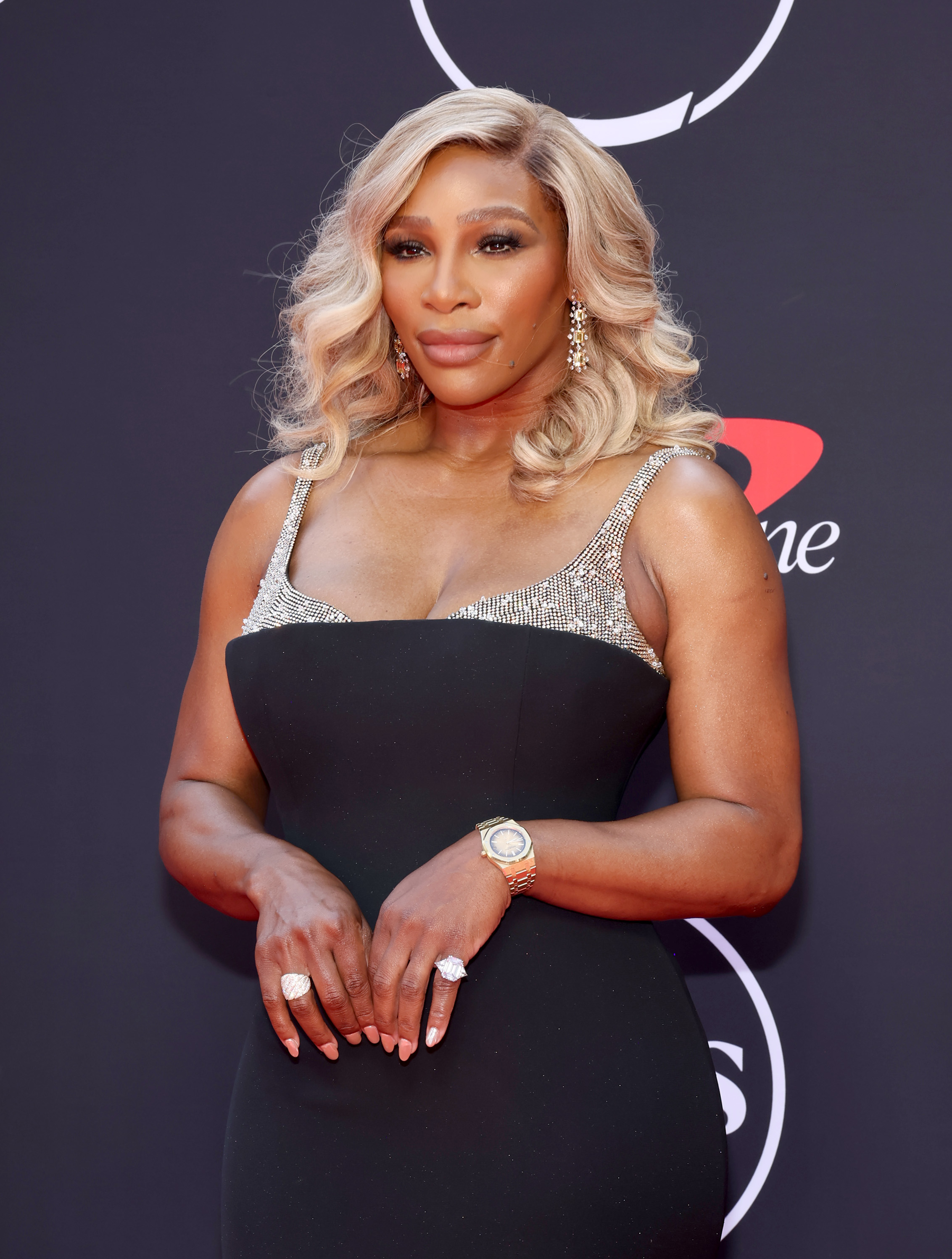 Serena Williams poses on the red carpet in an elegant black gown with silver accents, accessorized with statement jewelry and wavy hair