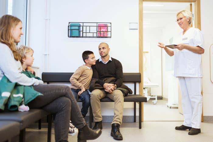 A nurse stands in a medical office doorway with a clipboard, speaking to a family seated in the waiting area: a man, two children, and a woman