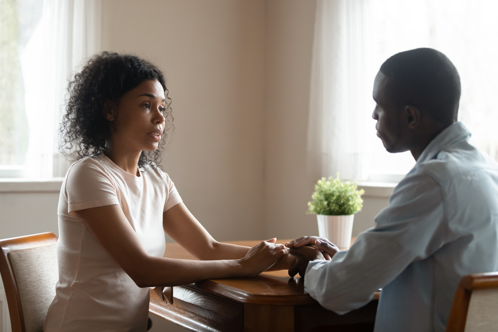 A woman and a man sit at a table facing each other, holding hands while having a serious conversation